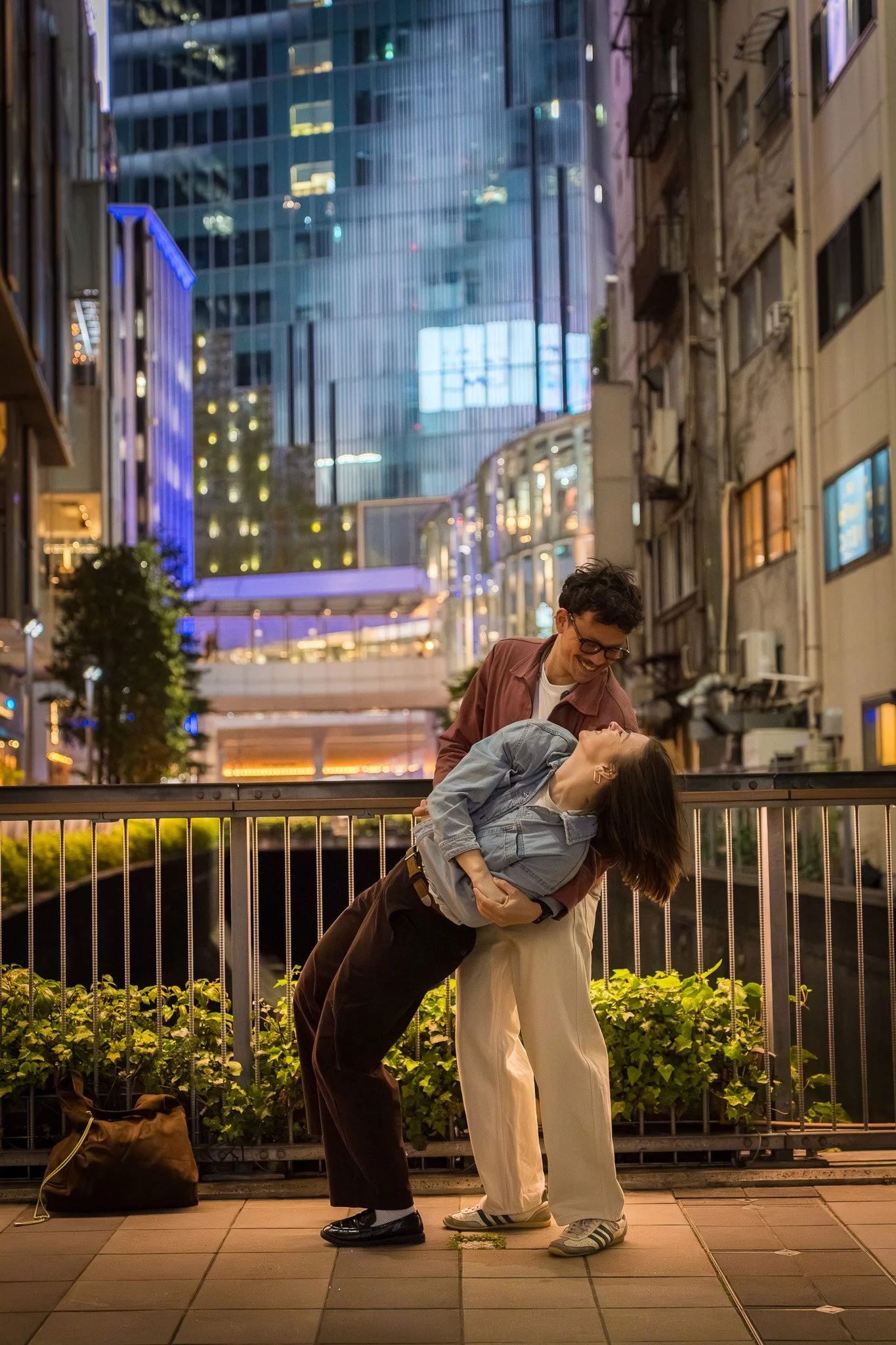 Two people, a man and a woman, are dancing and smiling on a city sidewalk at night with tall buildings and bright lights in the background.