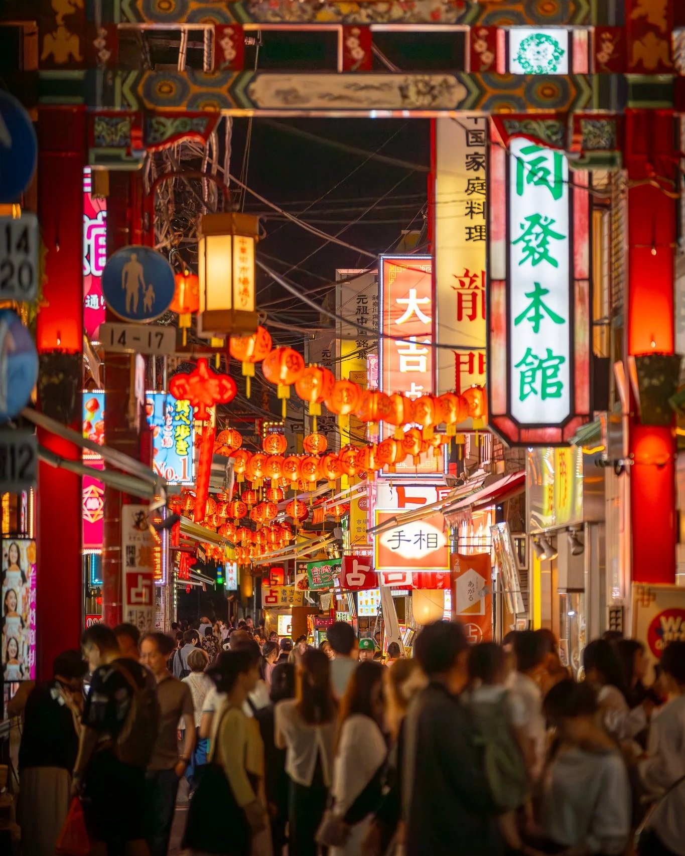 Crowded street scene at night in an Asian market with red lanterns, colorful signs, and many people shopping and walking, illuminated by streetlights and signage.