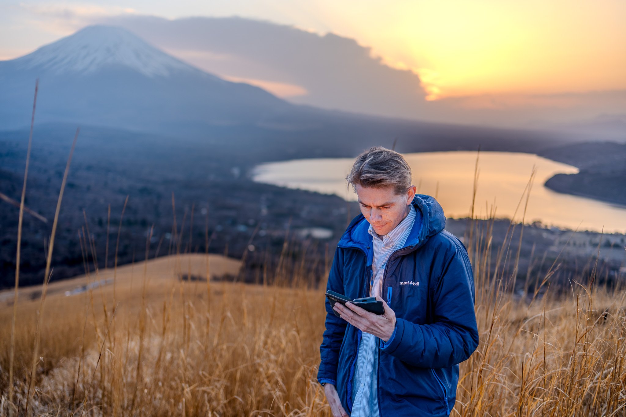 A man in a blue jacket holding a phone, standing in a grassy field with a lake, mountains, and a sunset in the background.