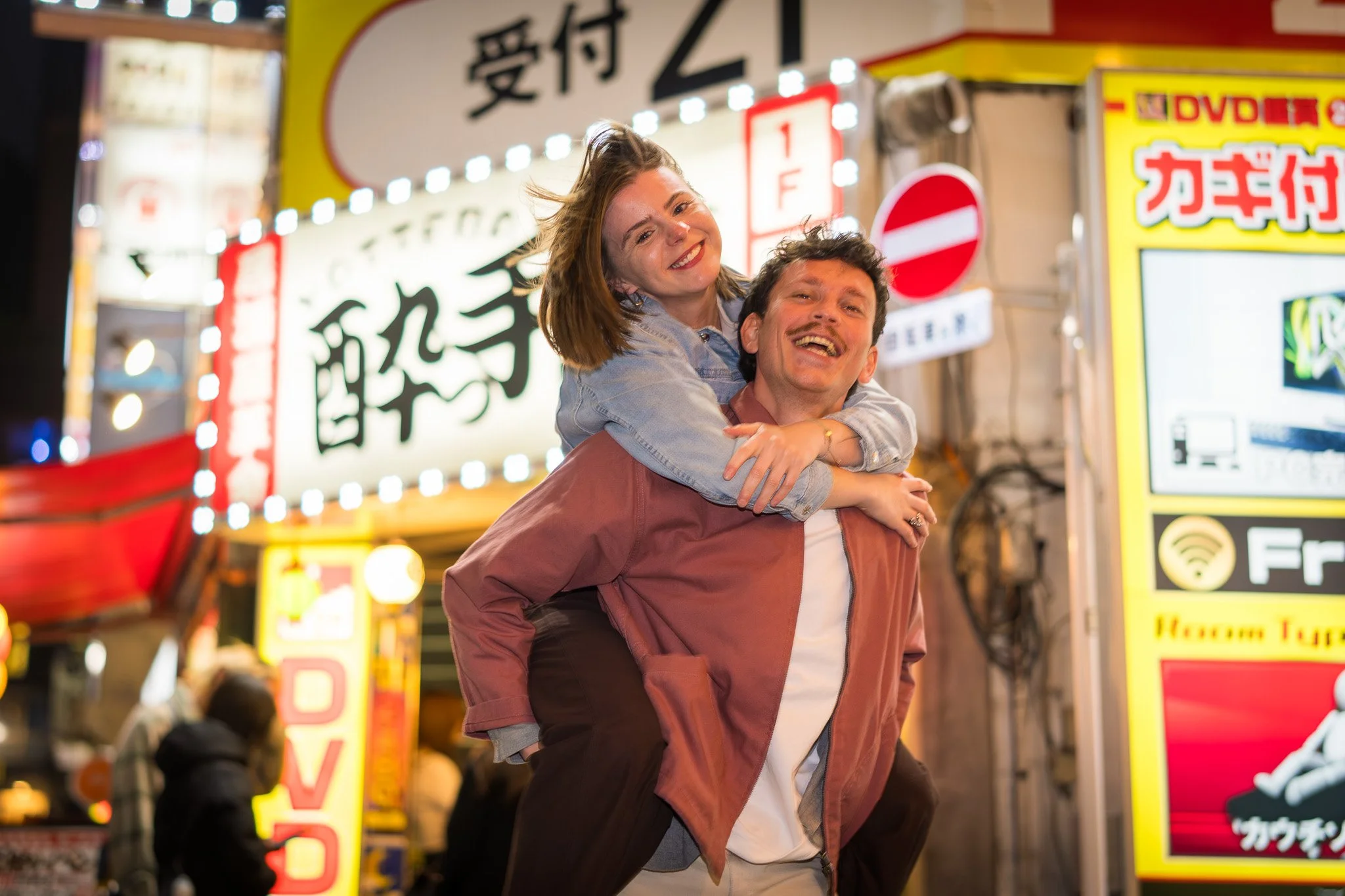A young woman and a man smiling and enjoying themselves in a busy, brightly lit street scene at night, with colorful signs in Japanese in the background.