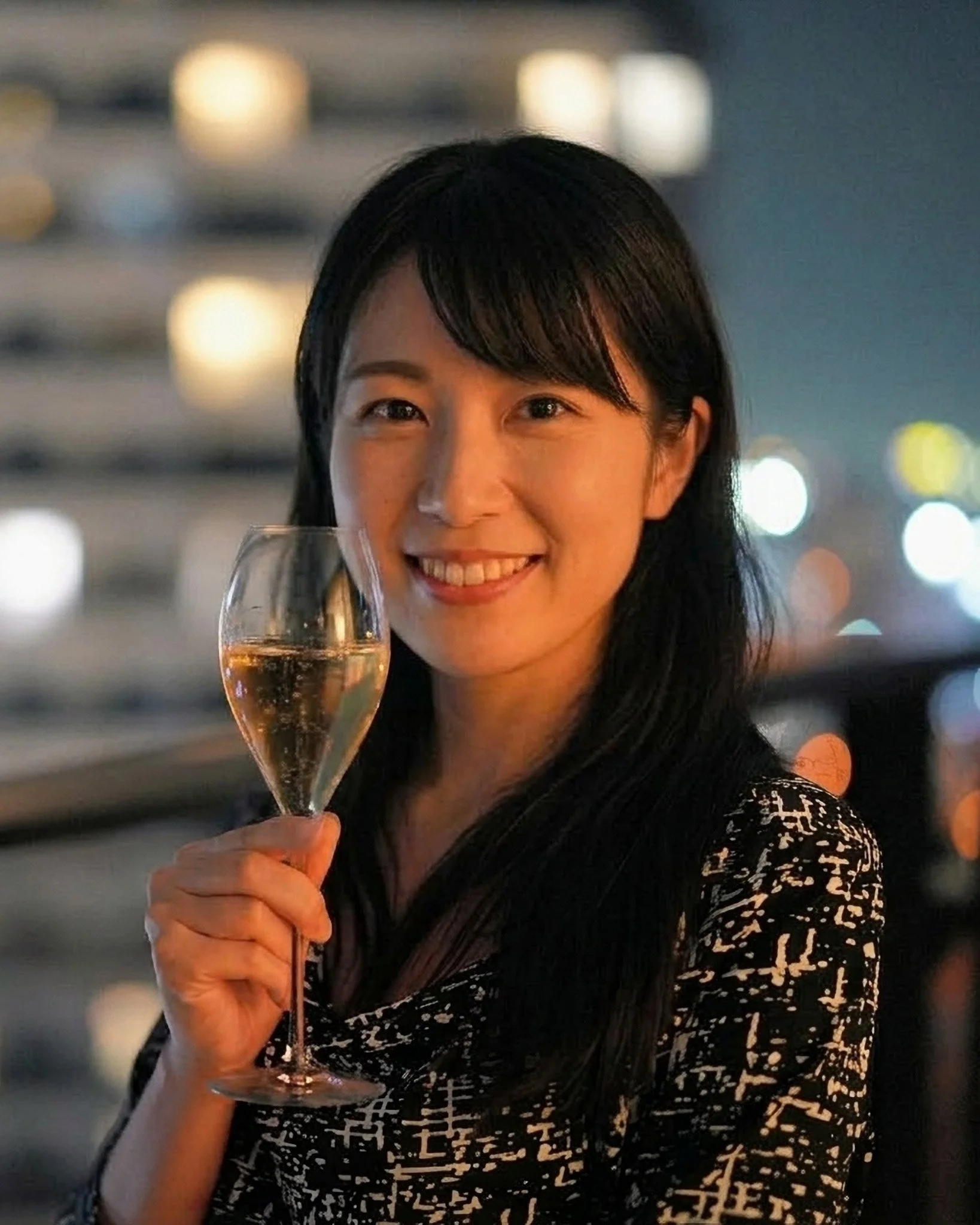 A woman smiling and holding a glass of champagne at night, with city lights in the background.