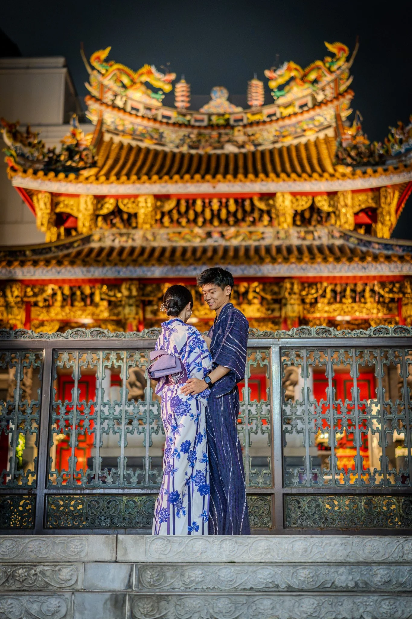 A couple dressed in traditional Japanese kimonos stands close together on a decorative stone staircase in front of a brightly lit ornate temple at night, with intricate architectural details and colorful decorations.