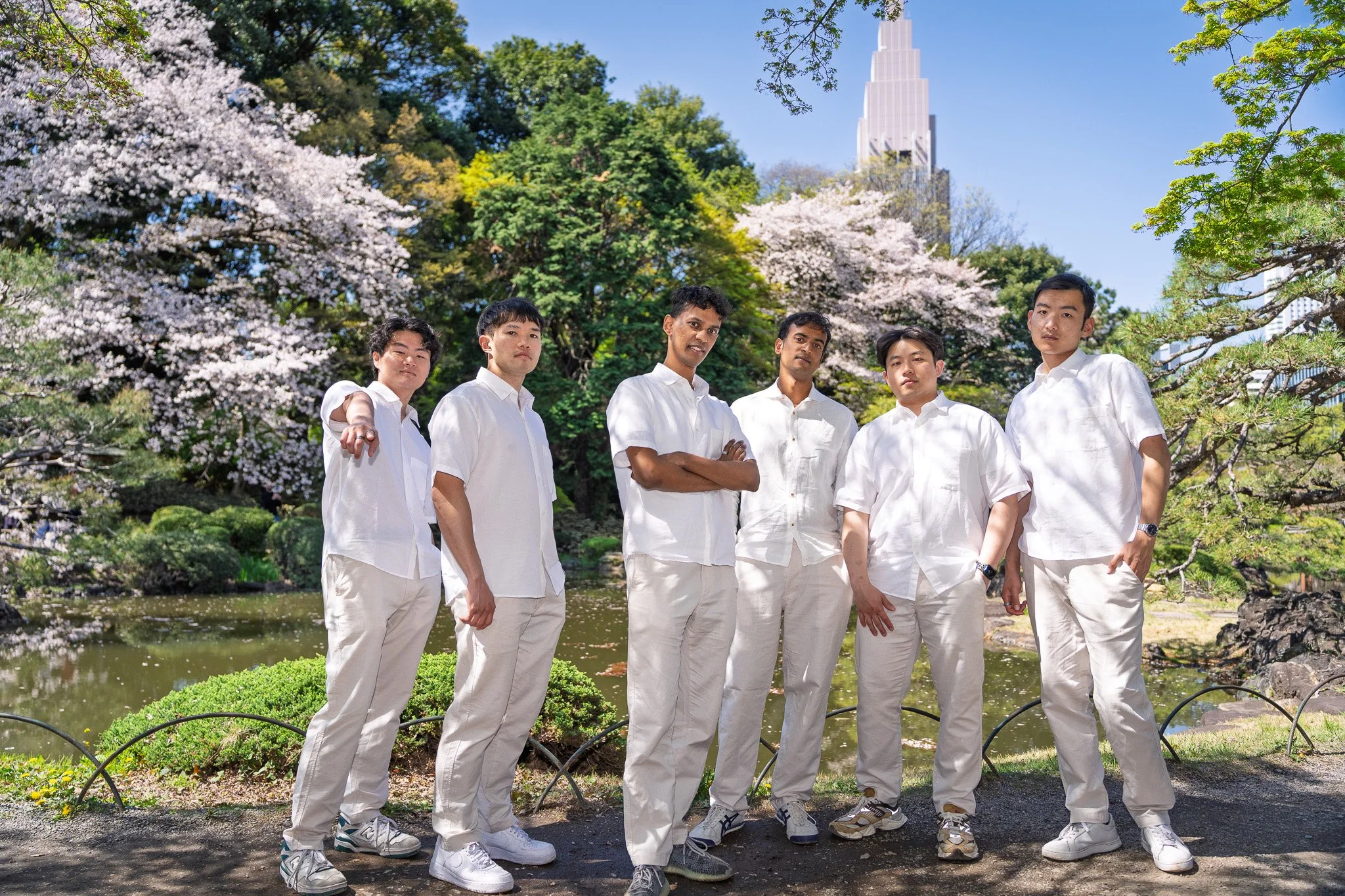 A group of six young men standing in a park with cherry blossom trees, a pond, and tall skyscrapers in the background. They are all dressed in white shirts and light-colored pants.
