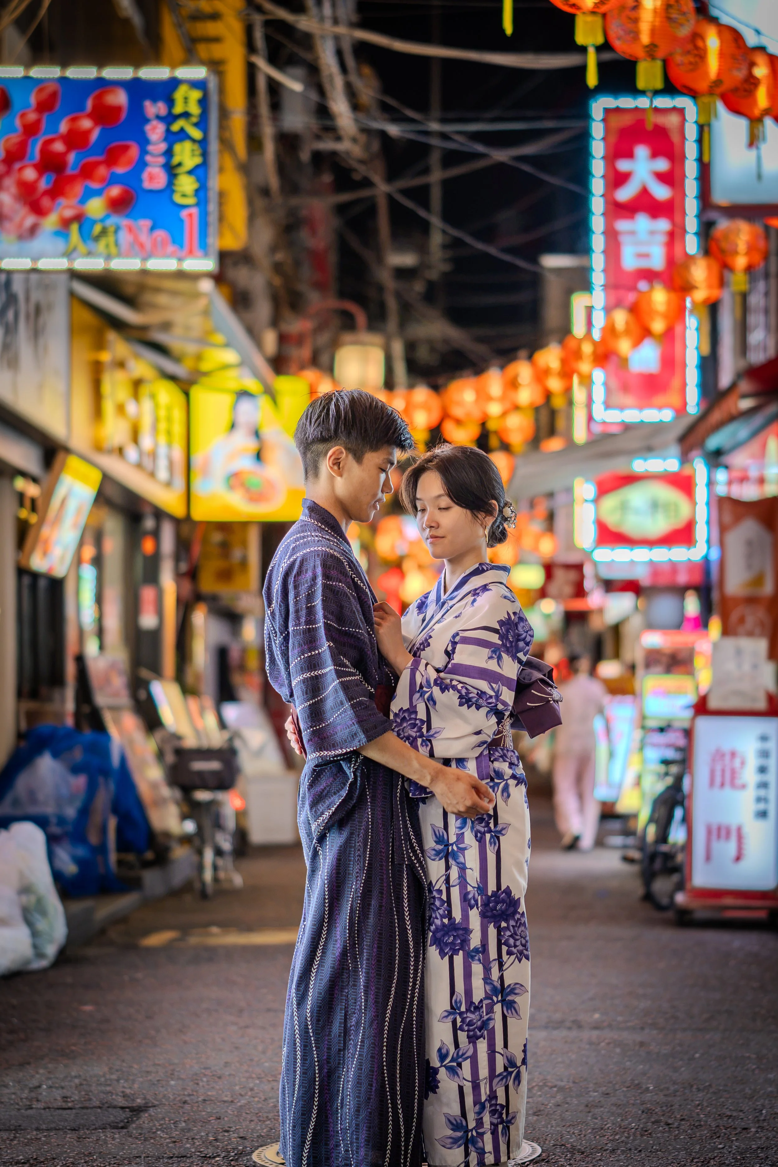 A young man and woman dressed in traditional Japanese kimonos stand close together at night in a lively, brightly lit street decorated with red lanterns and neon signs, sharing an intimate moment.