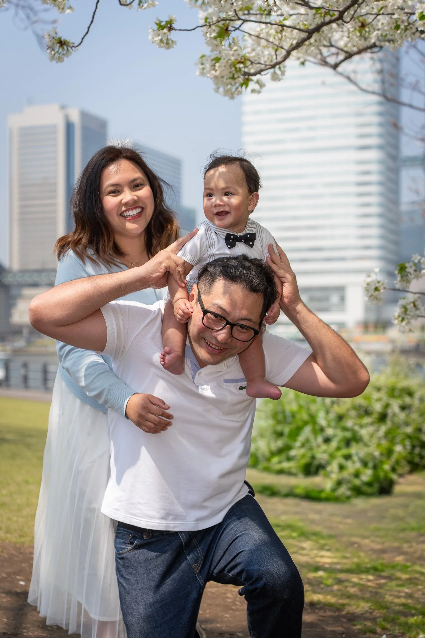 A family of three outdoors in a city park, with tall buildings in the background, smiling and playing together. The father carries the baby on his shoulders, the mother stands beside them, and the baby is wearing a striped shirt with a bow tie.