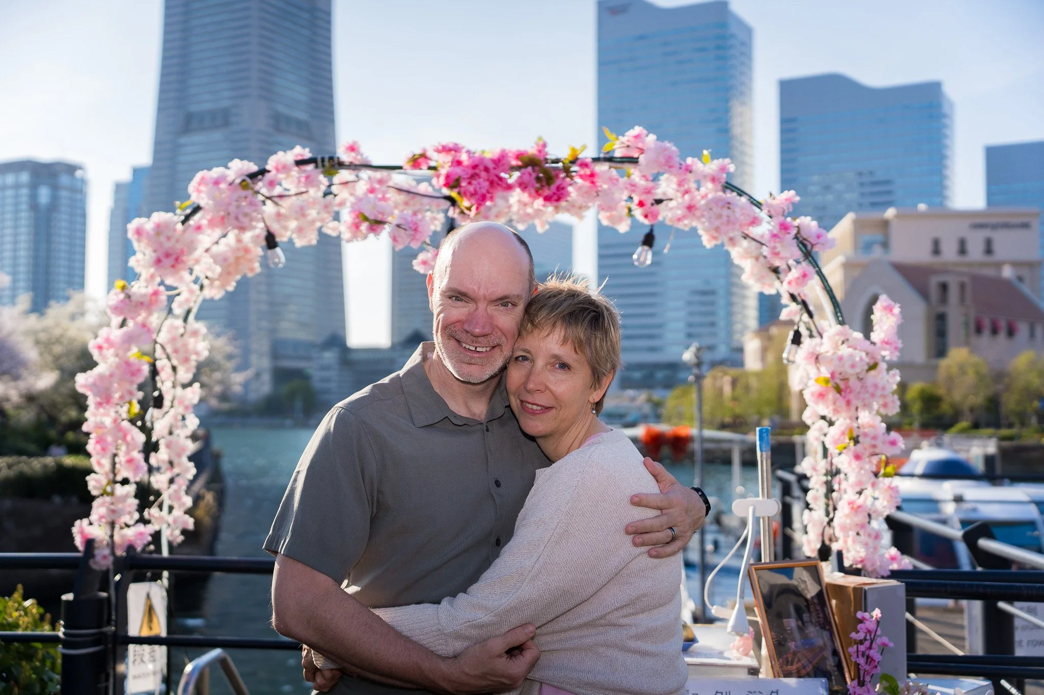 A smiling man and woman hugging in front of a floral arch near a waterway in an urban setting with high-rise buildings.