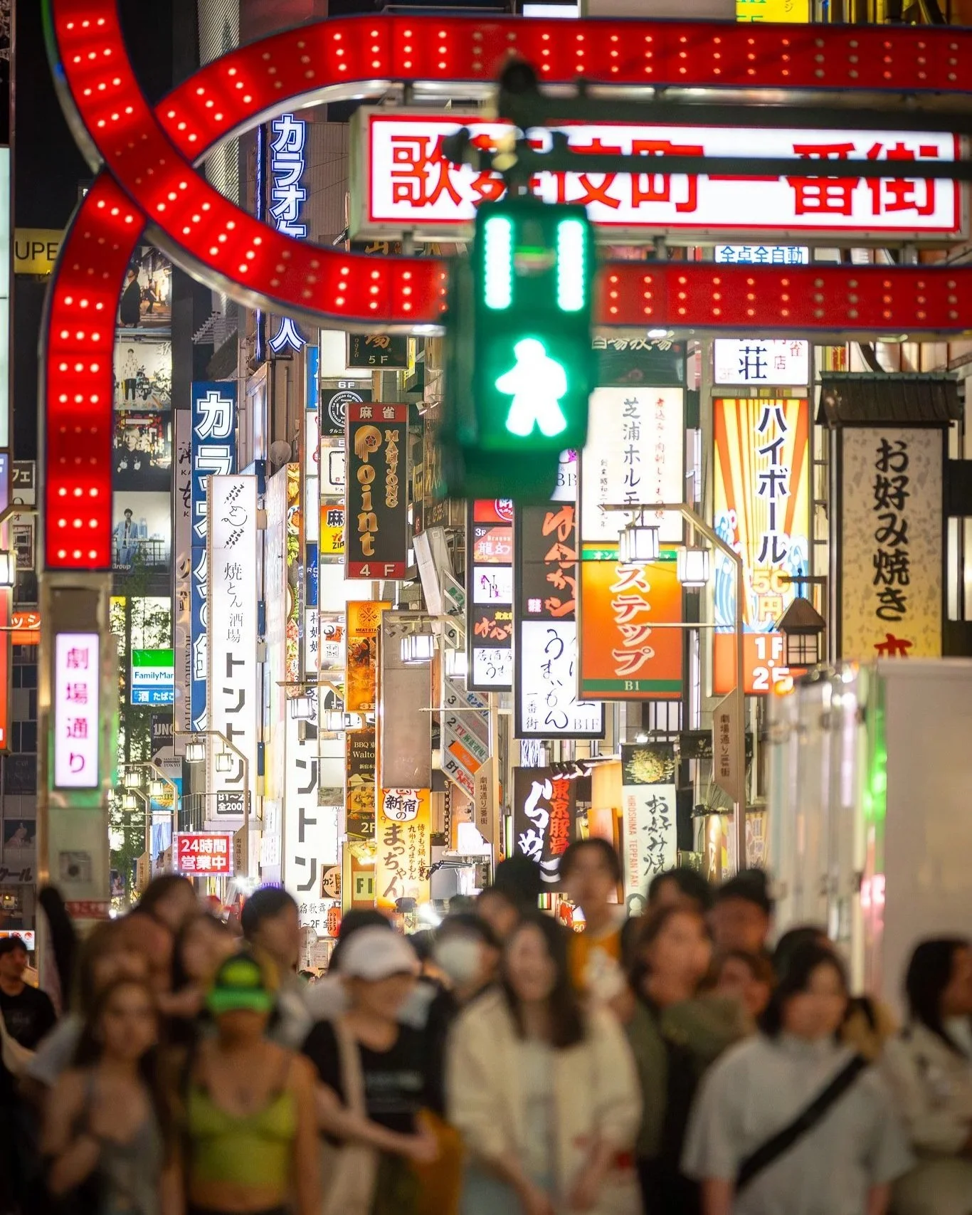 Nighttime crowded street in Japan with numerous neon signs and advertisements in Japanese, and a green pedestrian traffic light in the foreground.