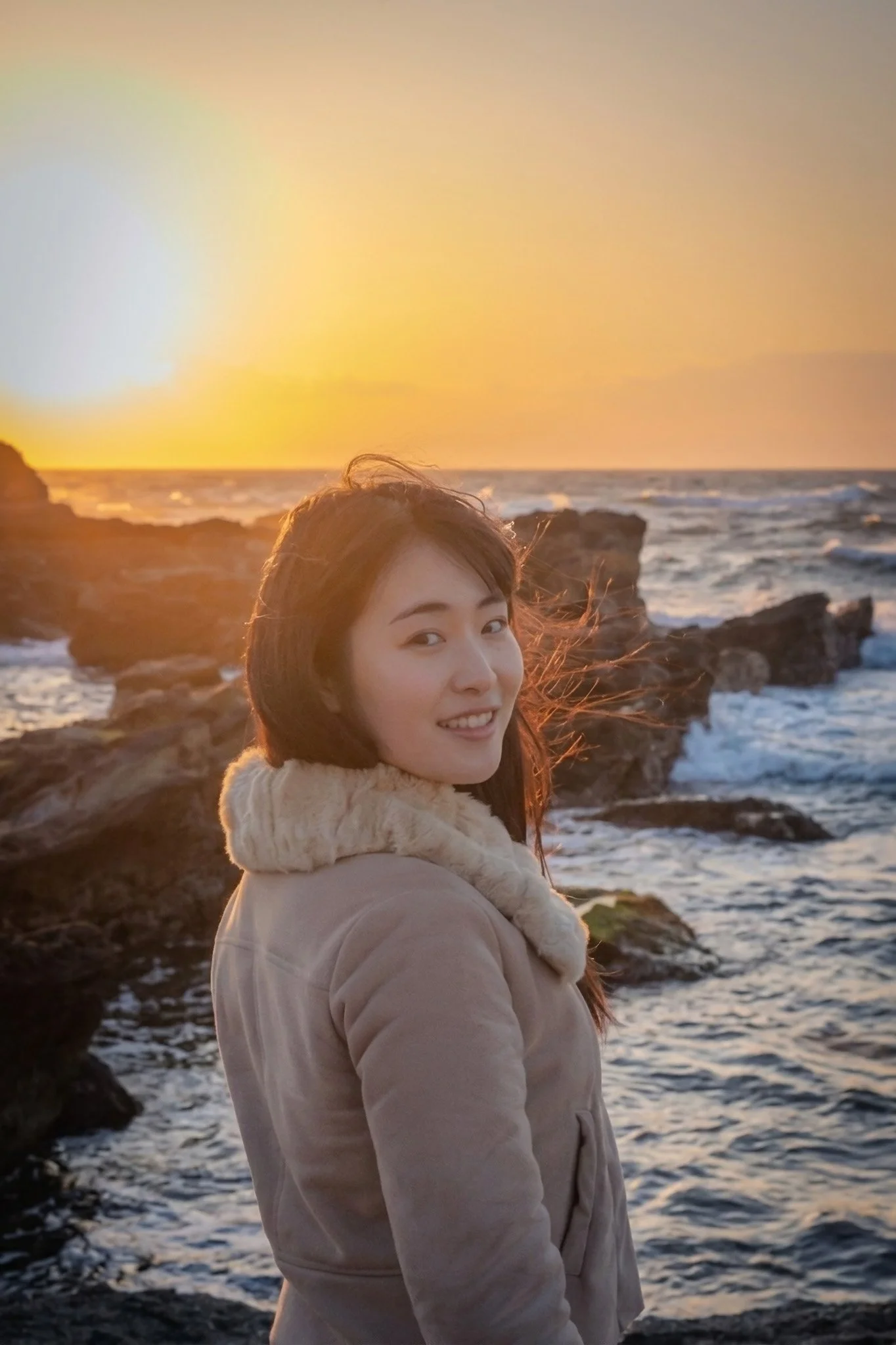 A woman smiling at the camera on a rocky coastline during sunset, with the ocean waves crashing behind her.