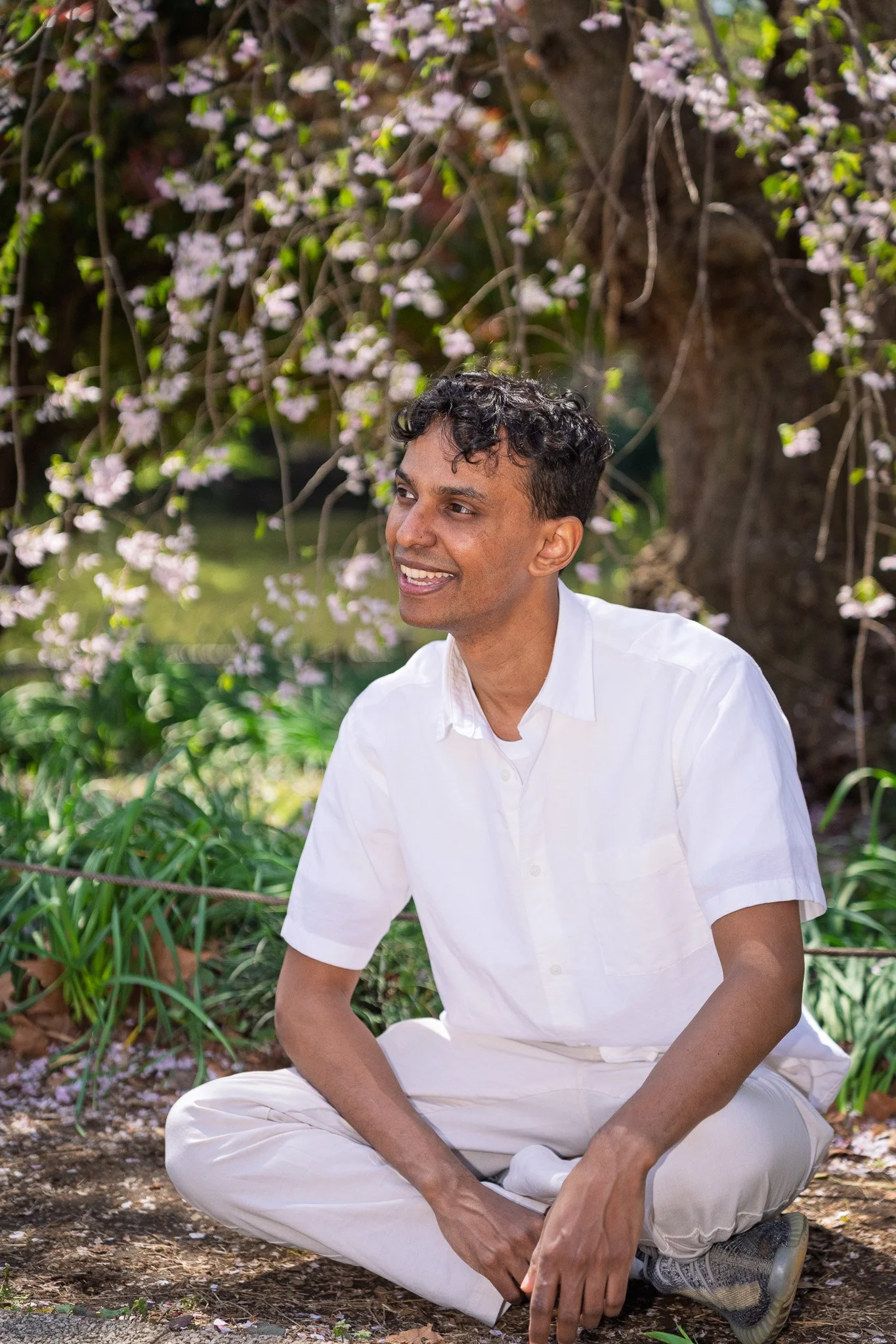 A man sitting cross-legged outdoors, smiling, with pink flowering tree and green foliage in the background.