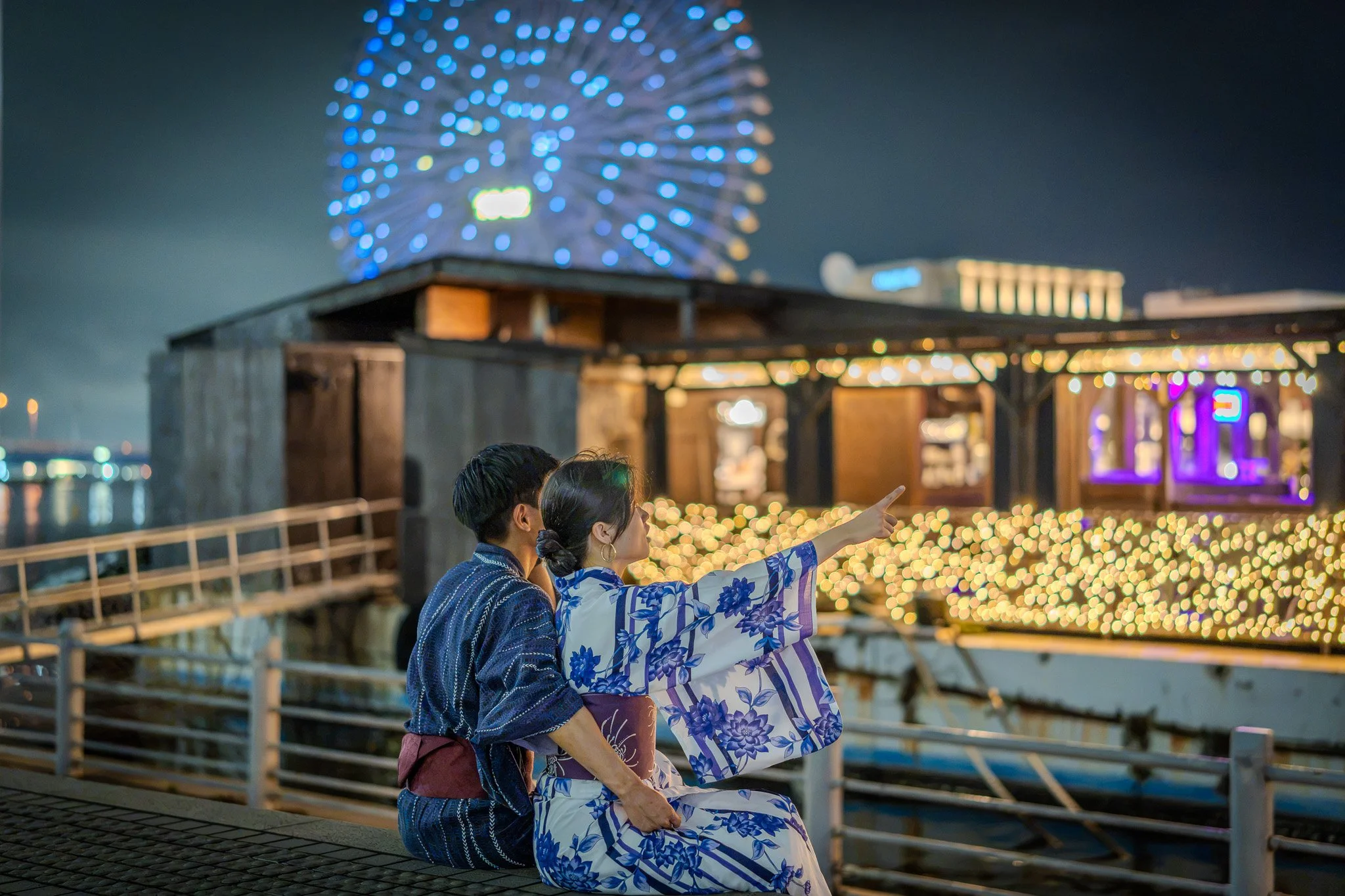 Couple in traditional Japanese kimono at night near a brightly lit Ferris wheel with colorful lights, with blurred festive lights in the background.