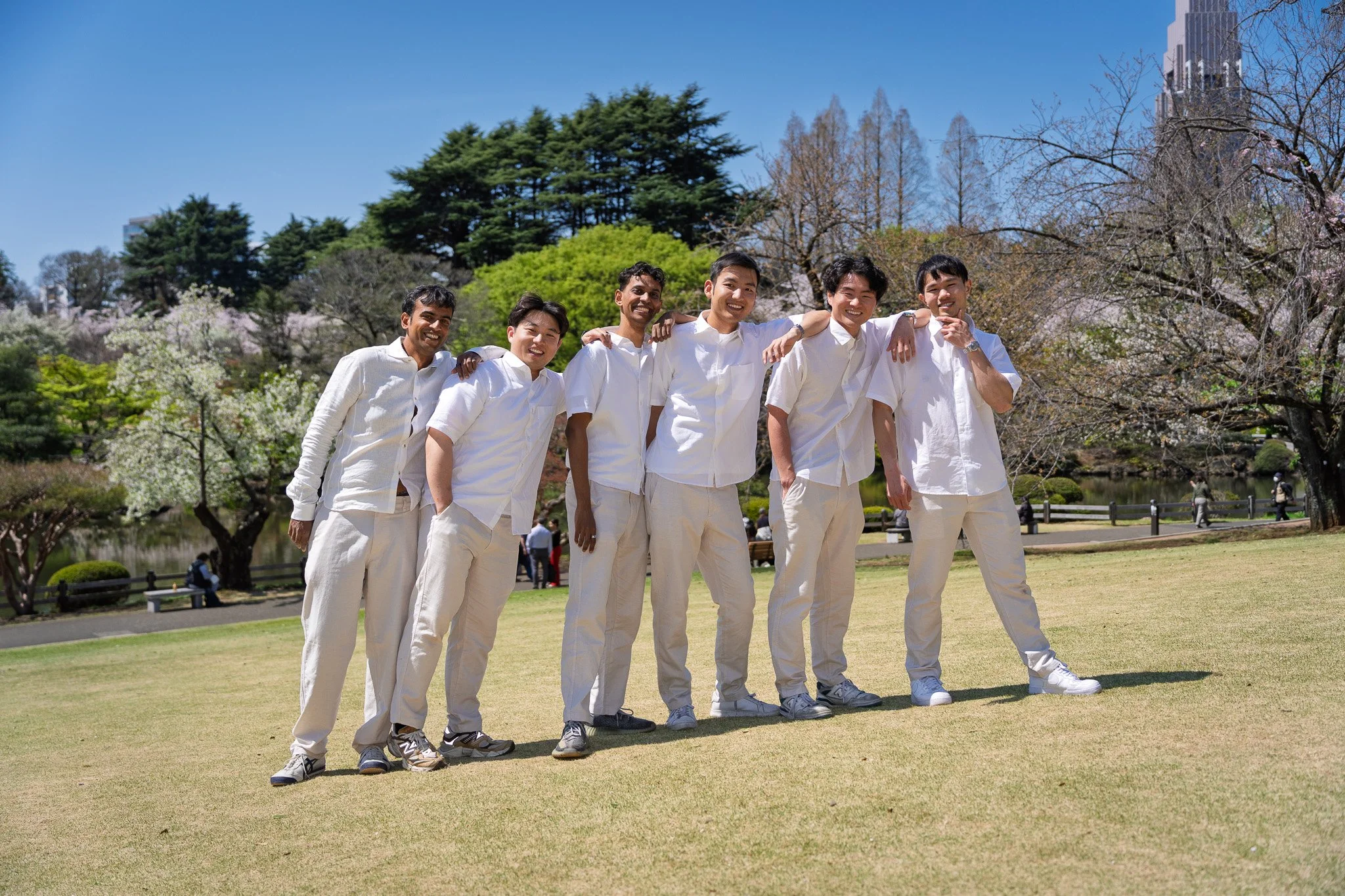 Group of six men standing closely together outdoors, smiling and posing for a photo in a park with trees and a pond in the background.
