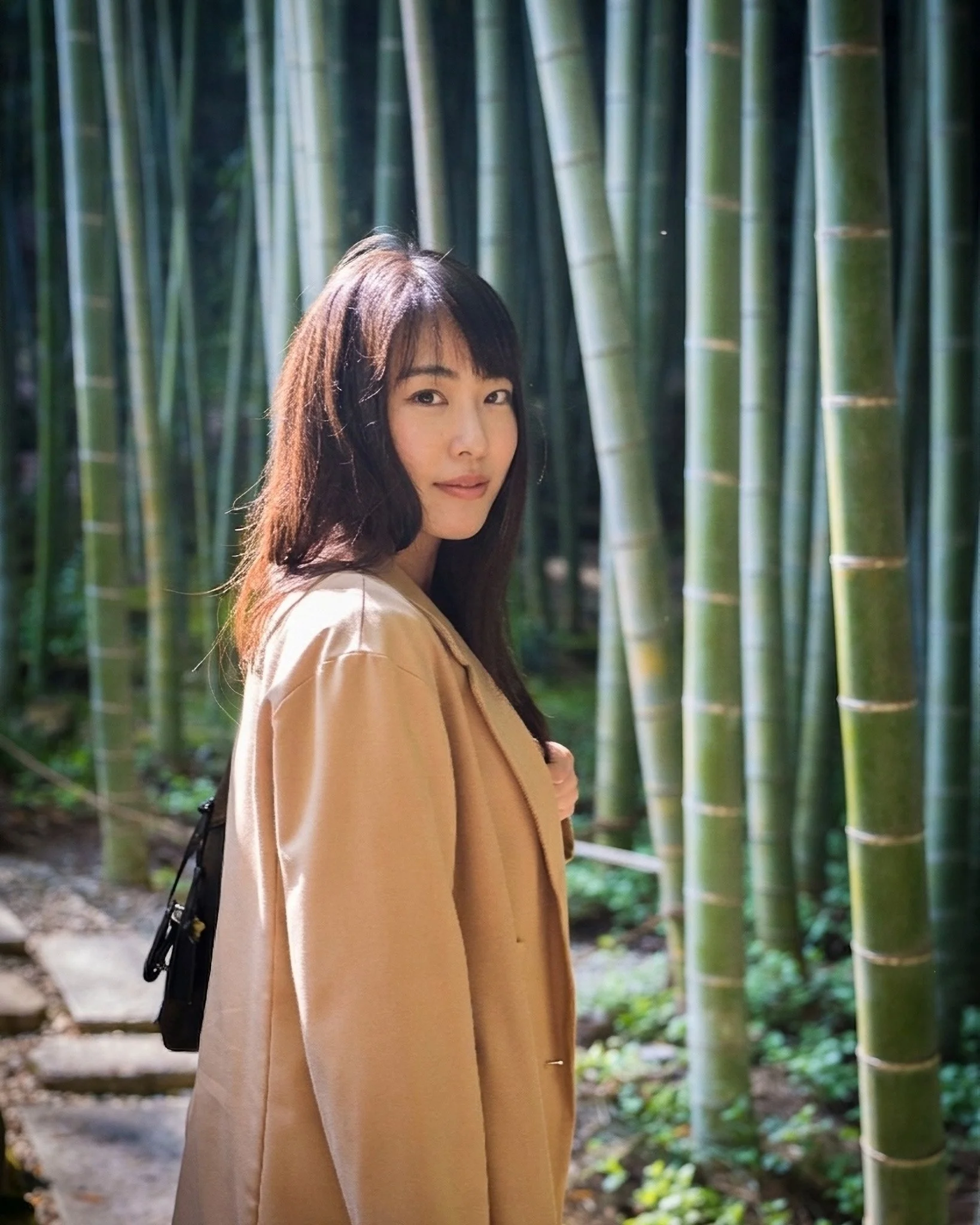 A woman standing in a bamboo forest, looking at the camera with a slight smile, wearing a beige coat and carrying a small black backpack.