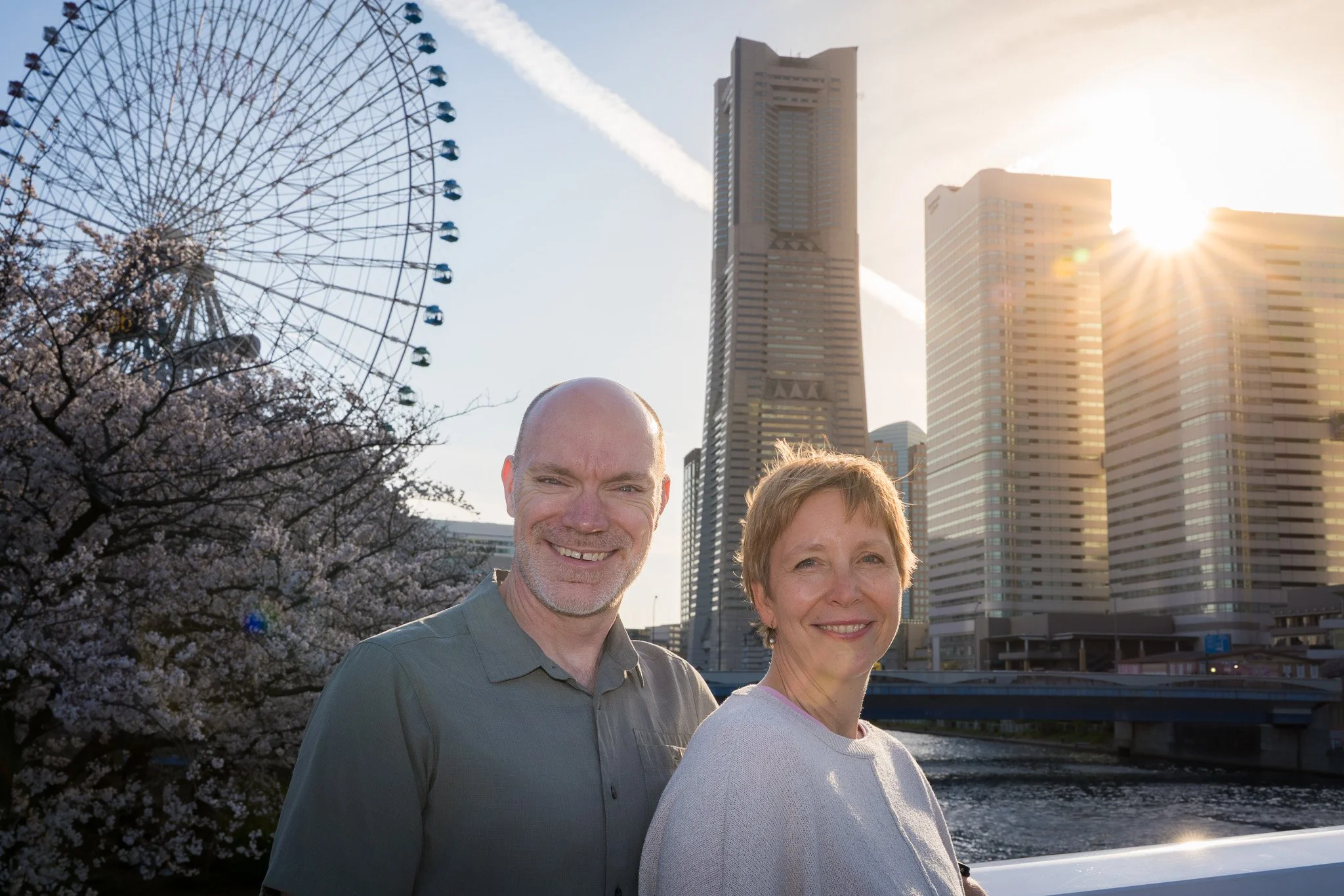 A smiling middle-aged man and woman standing outdoors near a river with city skyscrapers in the background during sunset, cherry blossoms visible on the left.