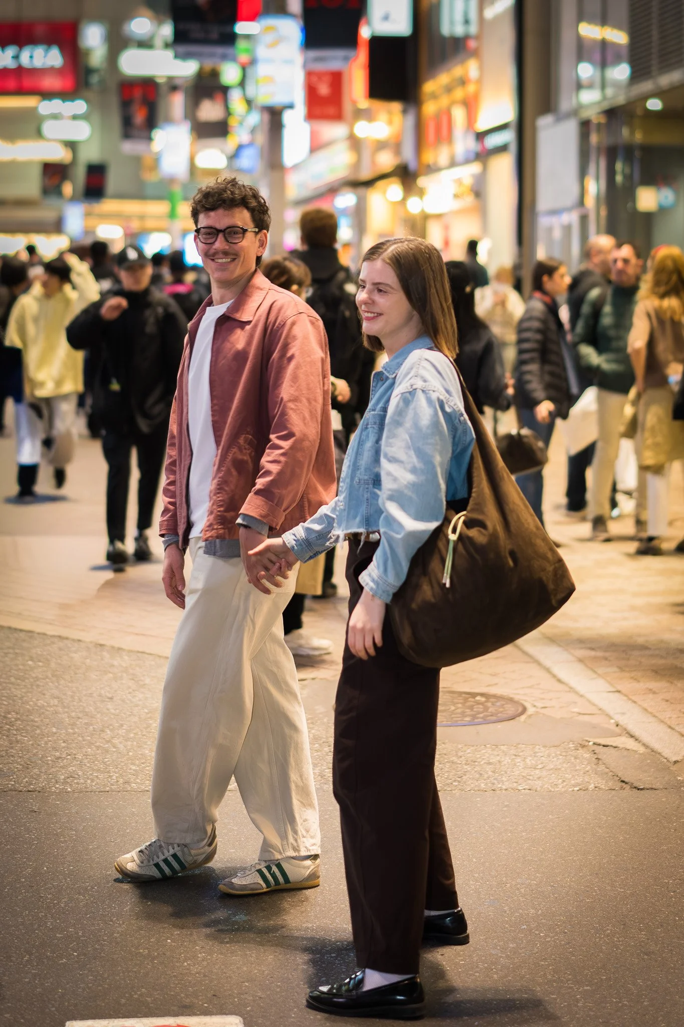 A young couple holding hands and smiling while crossing a busy city street at night, surrounded by other pedestrians and bright city lights.
