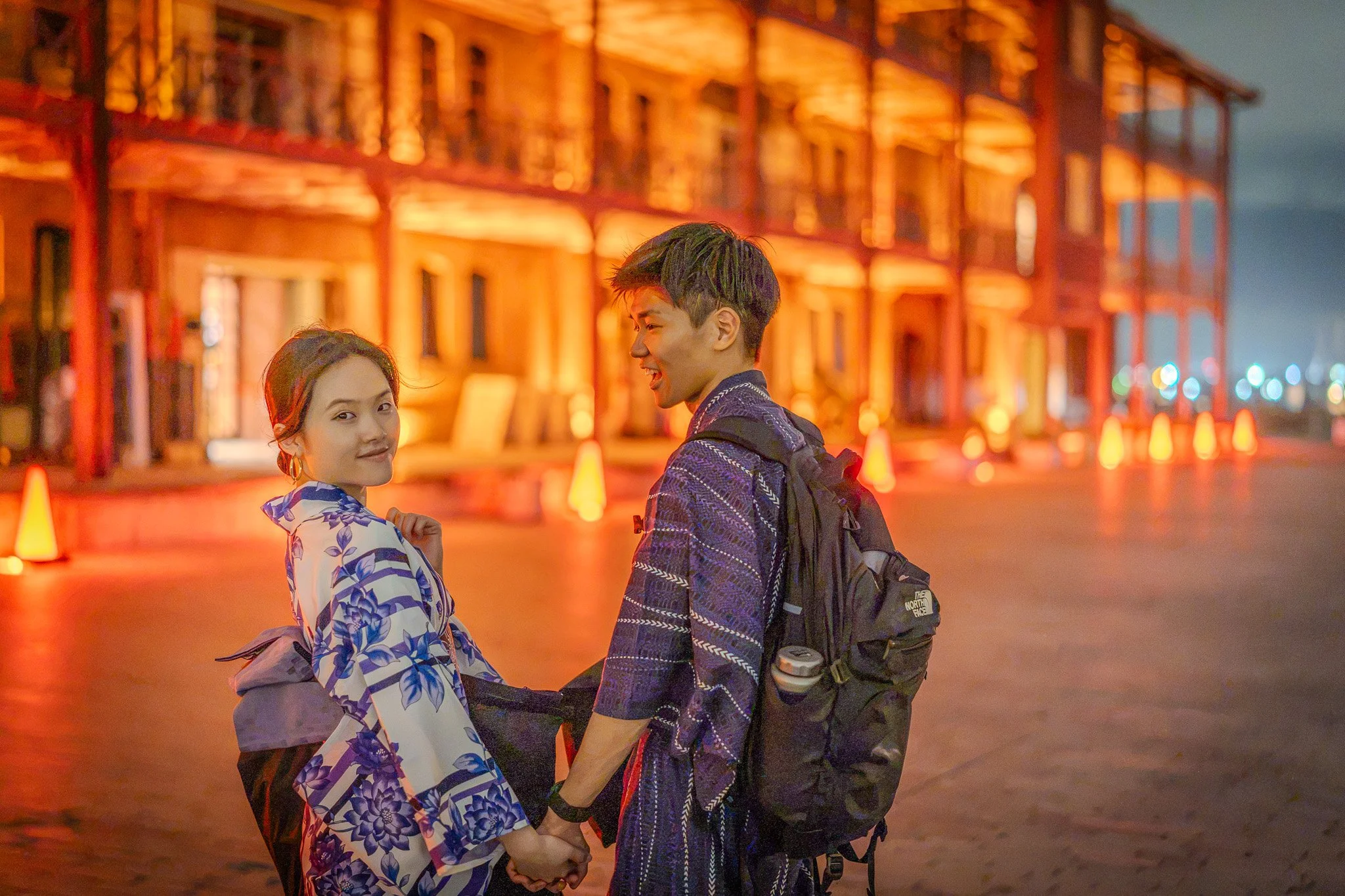 A young man and woman holding hands at night in front of a warmly lit building, smiling and looking at each other.
