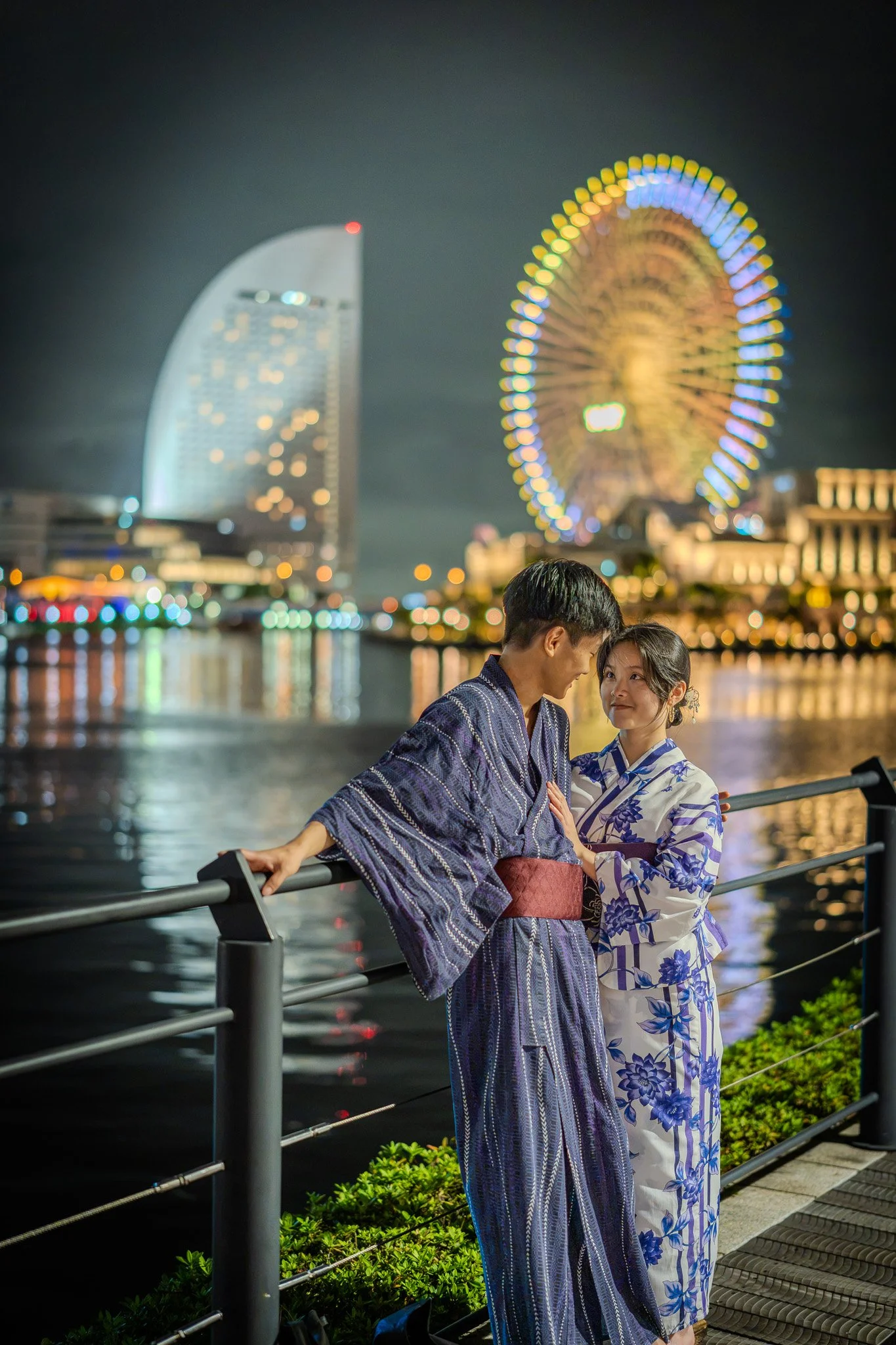 A couple dressed in traditional Japanese yukatas stands on a riverside at night, with a Ferris wheel and a modern city building illuminated in the background.