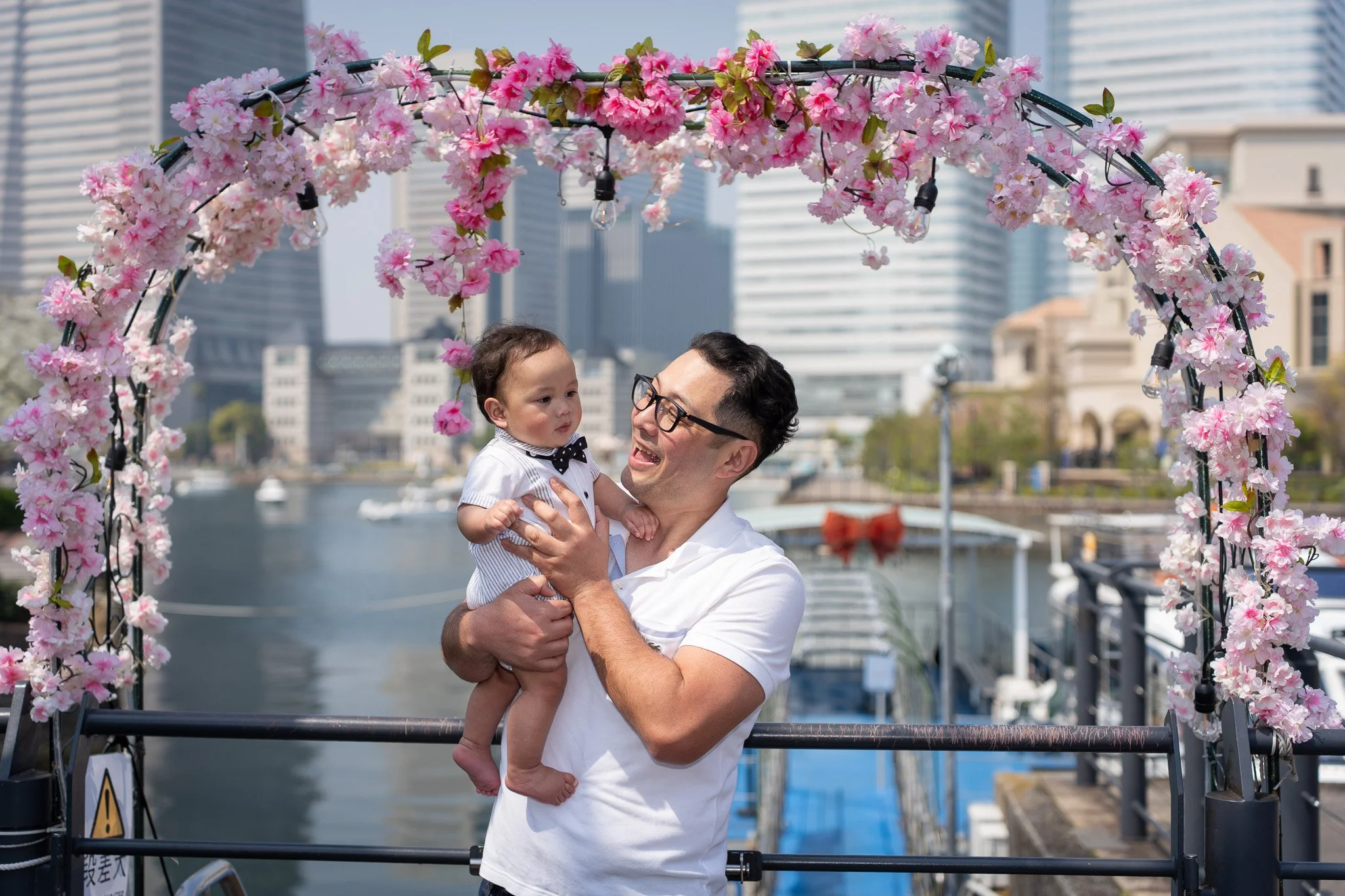 A man holding a young child under a pink cherry blossom arch near a waterfront with modern buildings and boats in the background.