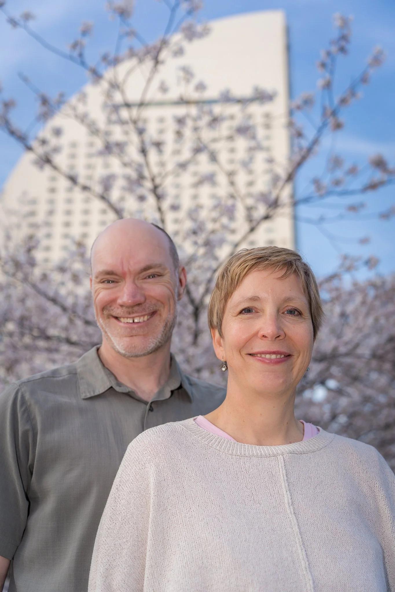 A smiling woman with short blonde hair and a man with short hair and beard pose outdoors in front of blooming cherry blossom trees and a tall building during daytime.