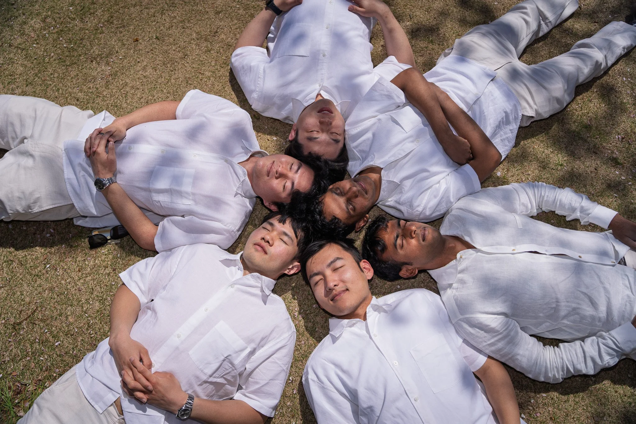 Group of seven adults lying on grass in white clothes, eyes closed, forming a star shape, outdoors, sunny day.