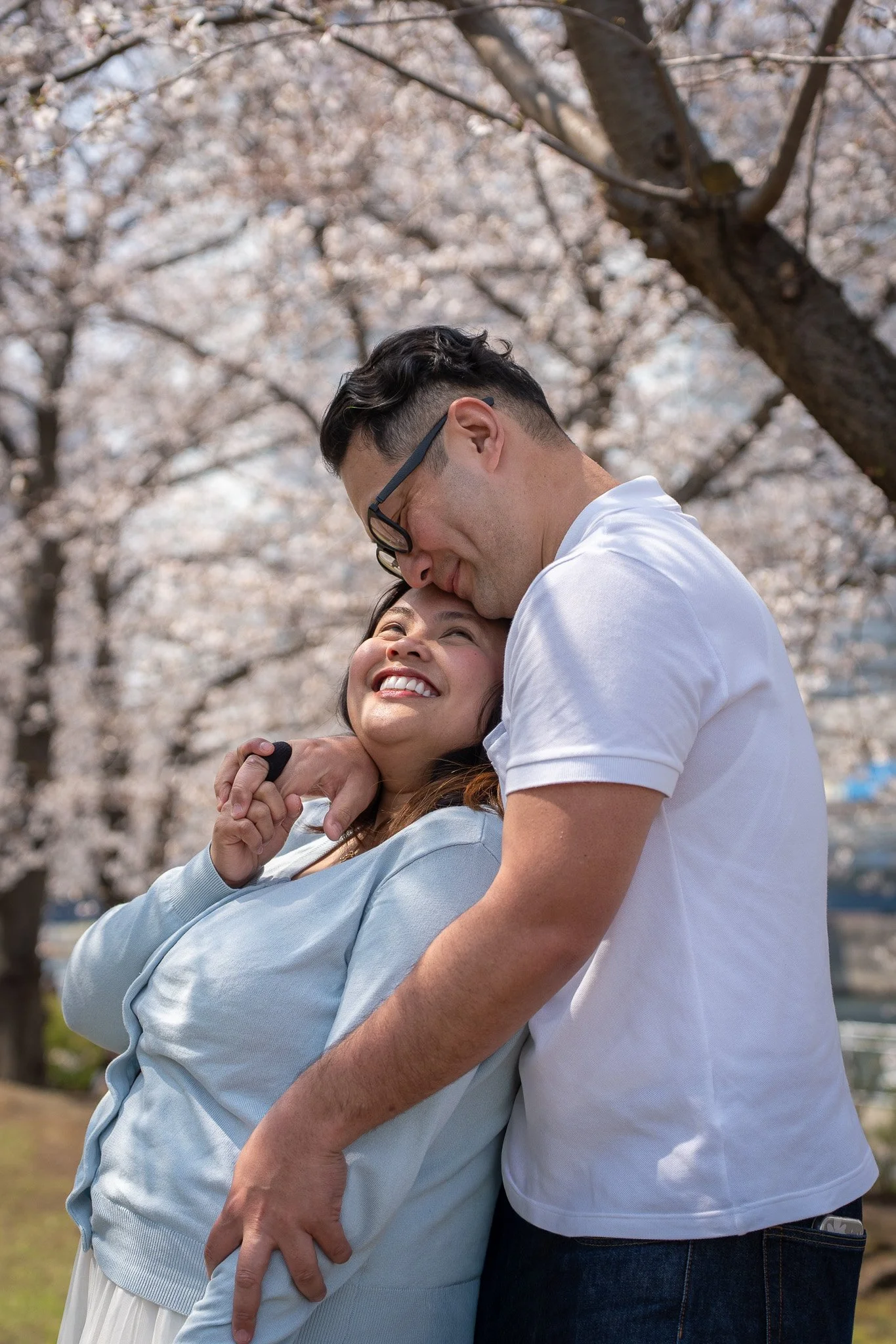 A couple in a loving embrace, smiling and looking at each other, standing under blooming cherry blossom trees.