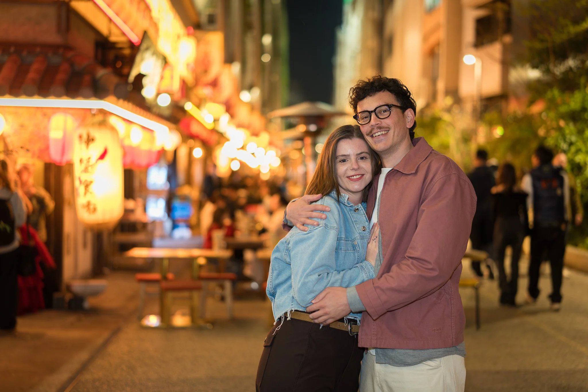 A young couple smiling and hugging outdoors at night, with colorful lights and blurry people in the background.