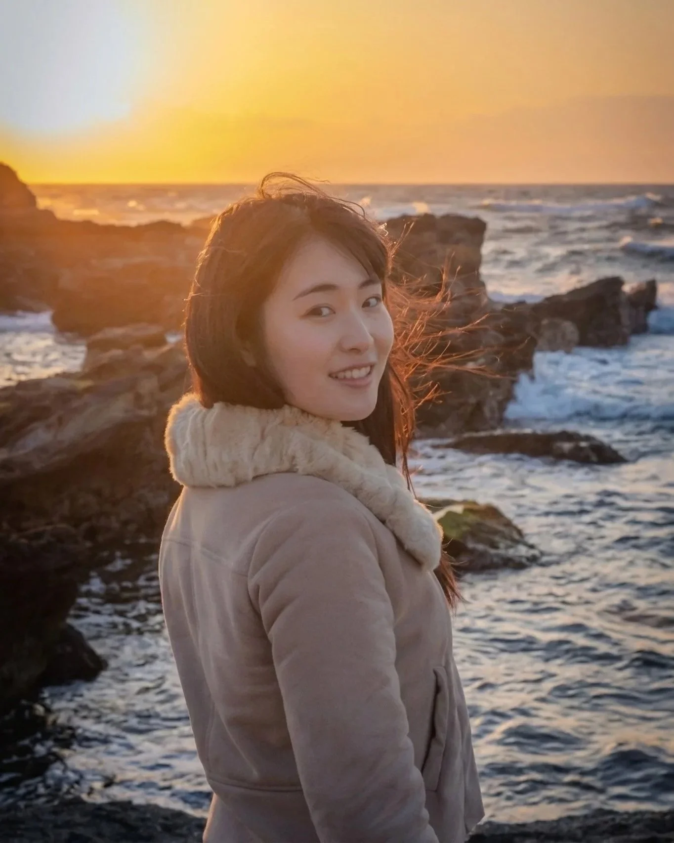 A woman with dark hair wearing a beige jacket with a faux fur collar stands by the rocky shoreline at sunset, smiling at the camera with the ocean and setting sun in the background.