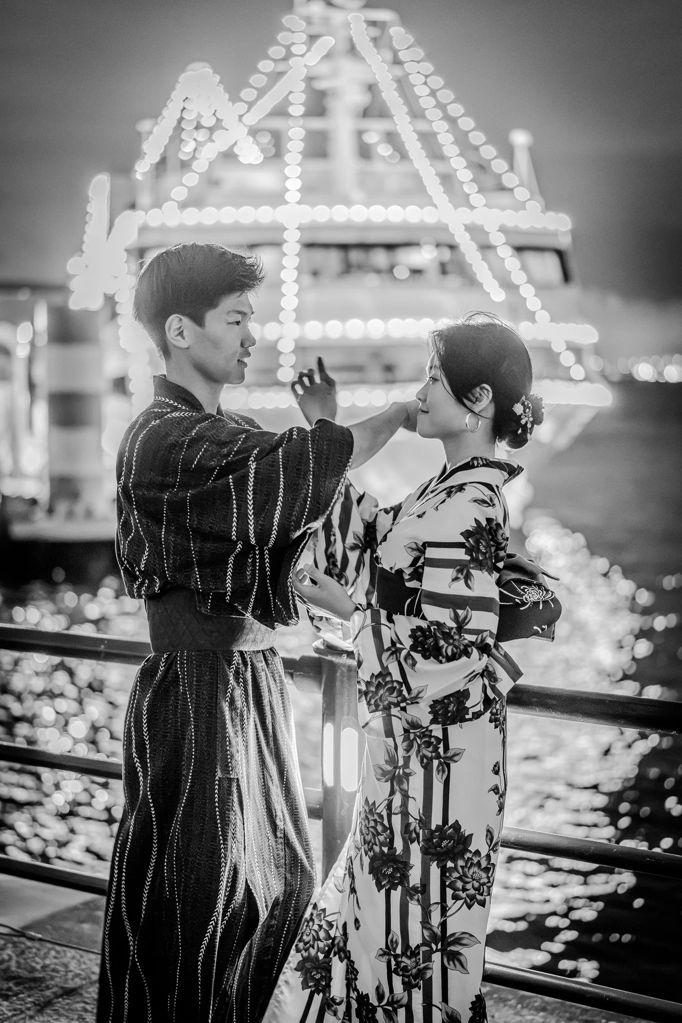 A black-and-white photo of a man and woman in traditional Japanese kimonos standing close to each other at a night fairground, with a decorated boat illuminated with lights in the background.