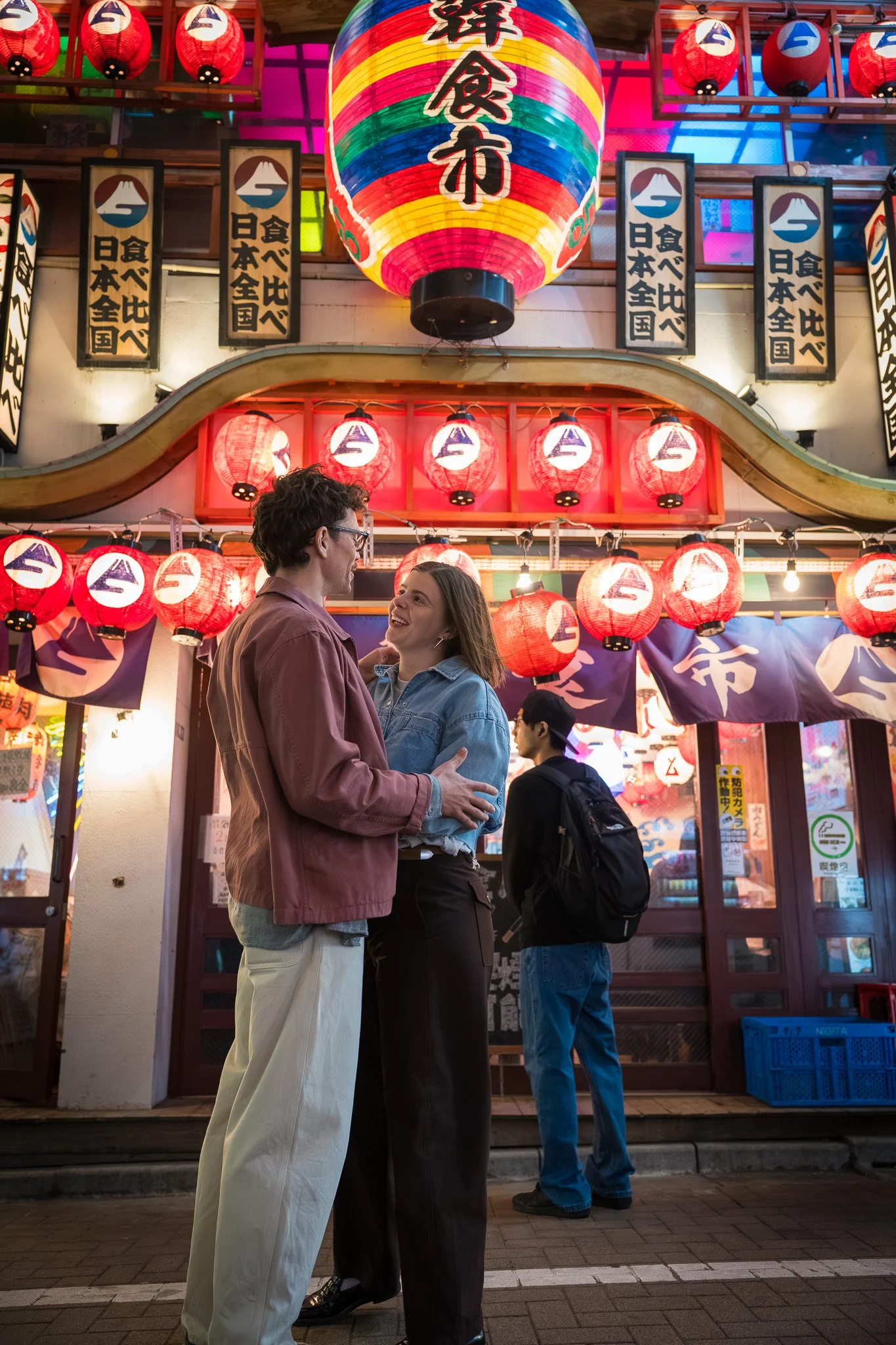 A couple smiling and talking in front of a colorful illuminated Japanese street market at night, decorated with red paper lanterns and signs.