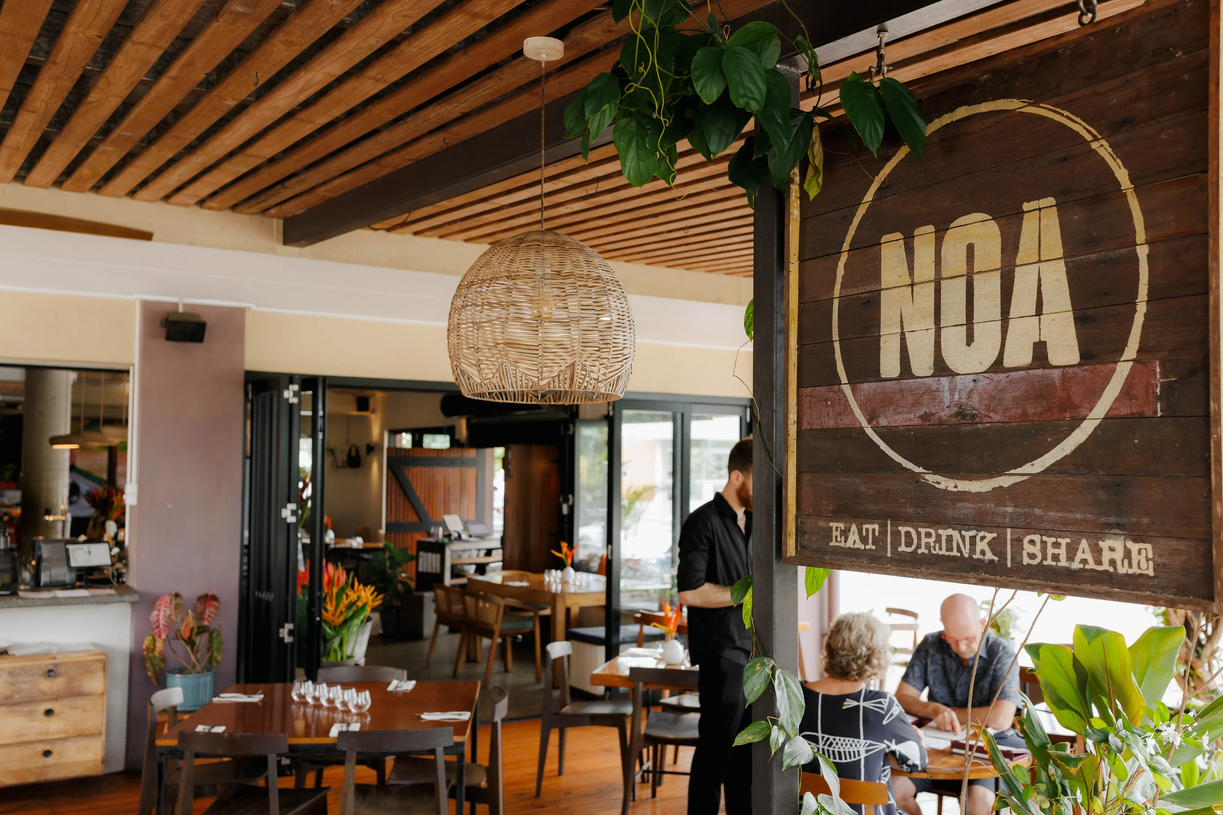 Interior of a restaurant or cafe with wooden and natural decor elements, including tables, chairs, and a hanging wicker lamp. A sign with the word 'NOA' and the words 'Eat | Drink | Share' is visible, with glimpses of customers seated at tables and a