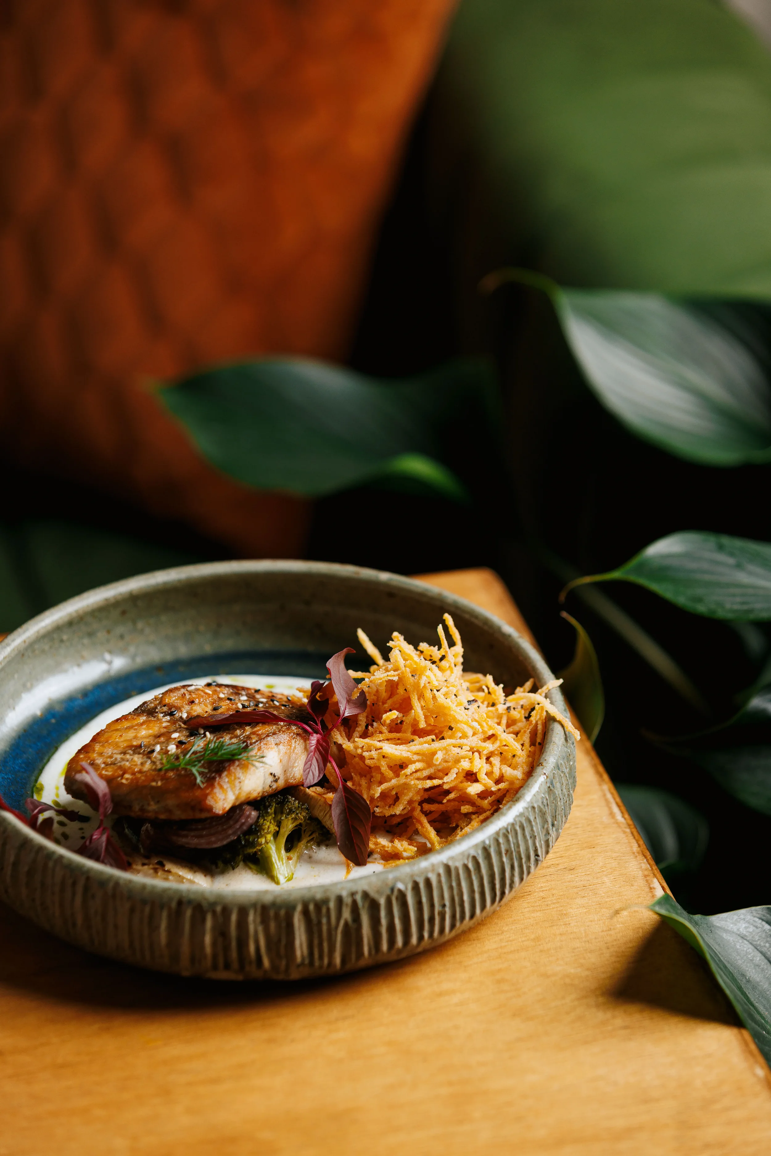 A ceramic bowl containing cooked fish, shredded vegetables, and garnishes, placed on a wooden surface with green leafy plants in the background.