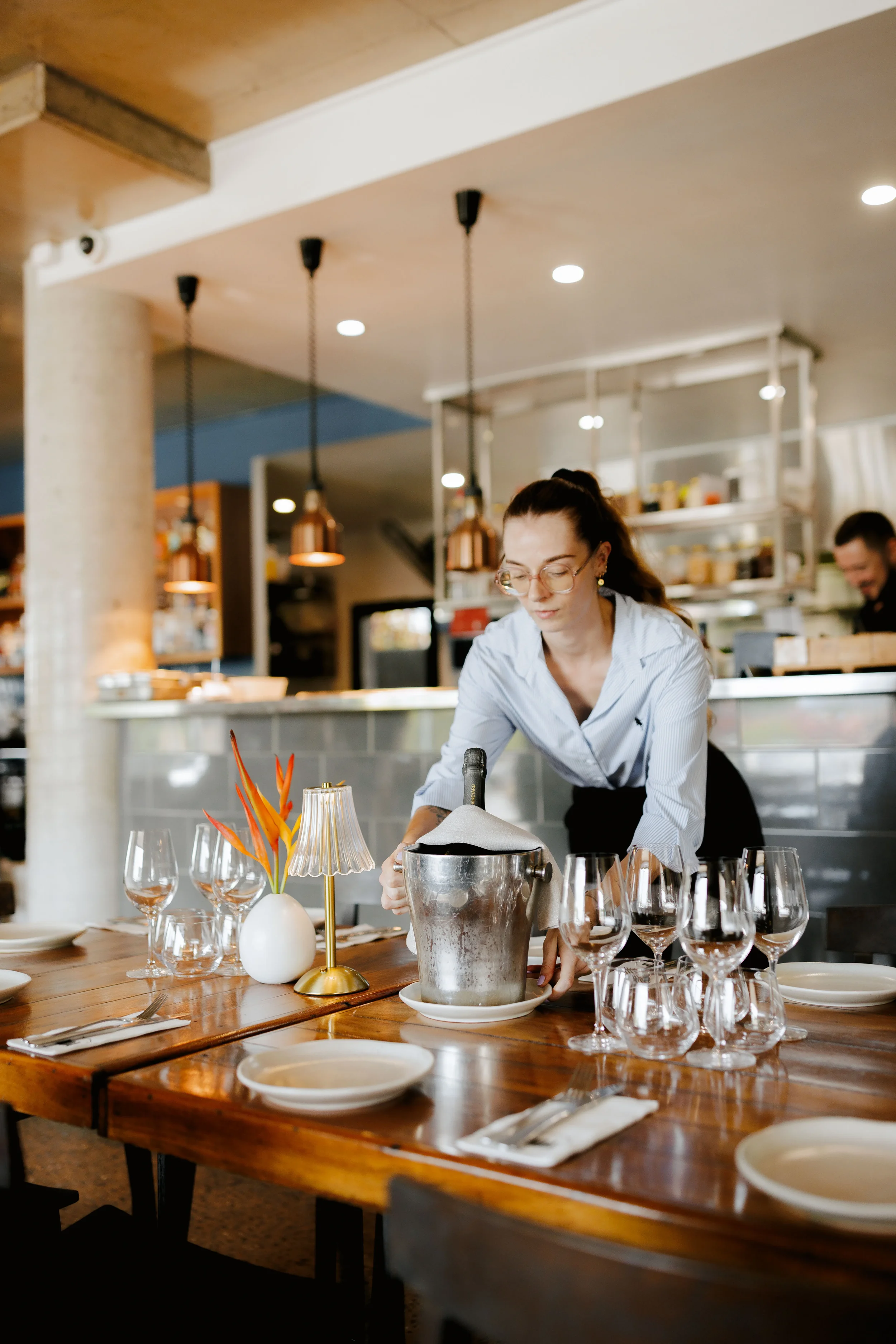 A woman in a light blue shirt is pouring champagne into glasses on a wooden table set for a meal, with a small lamp and a vase with orange flowers decoratively arranged.