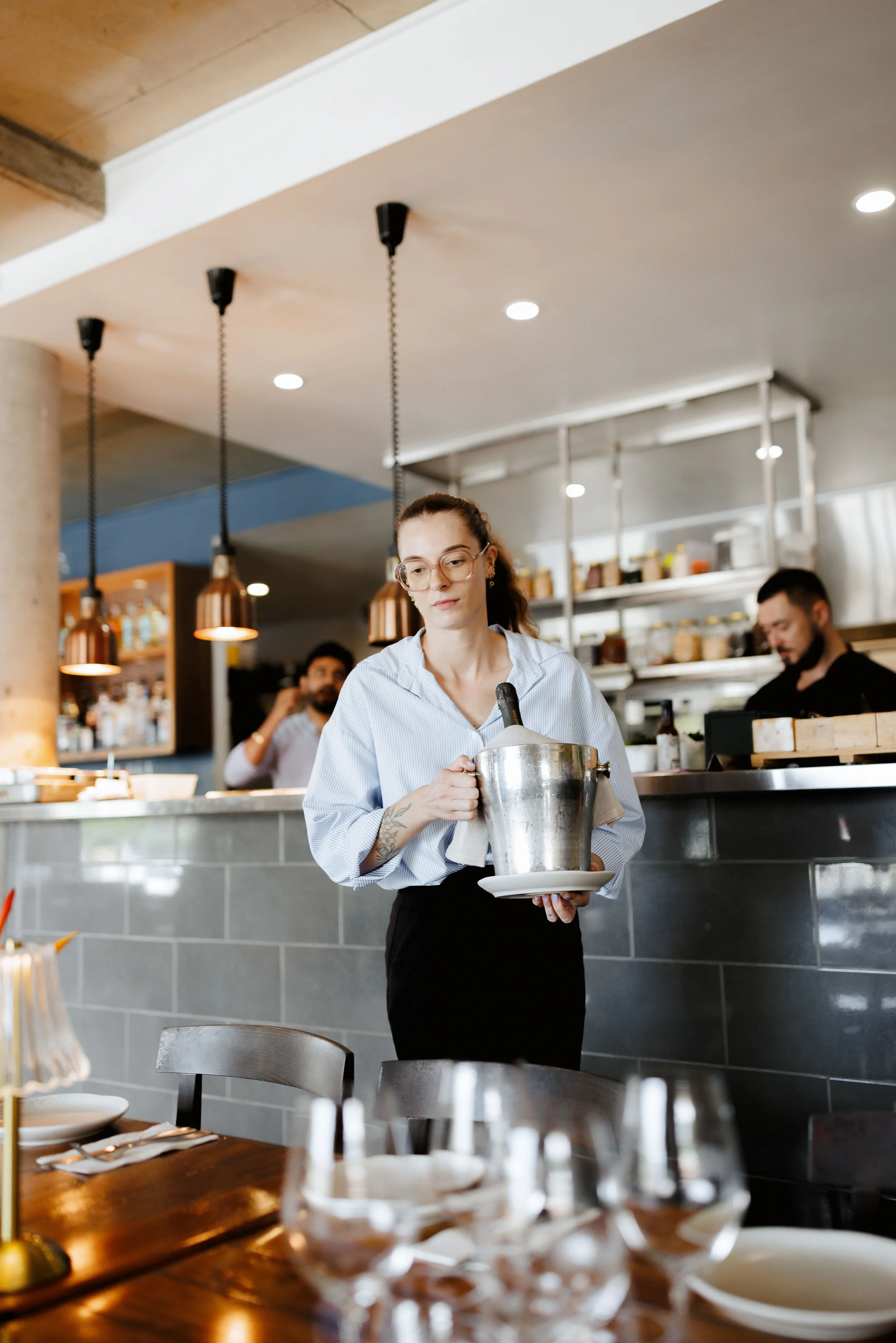 A waitress with glasses and a ponytail holding a plate and an ice bucket in a restaurant with a gray tile bar and shelves, pendant lights, and other staff in the background.