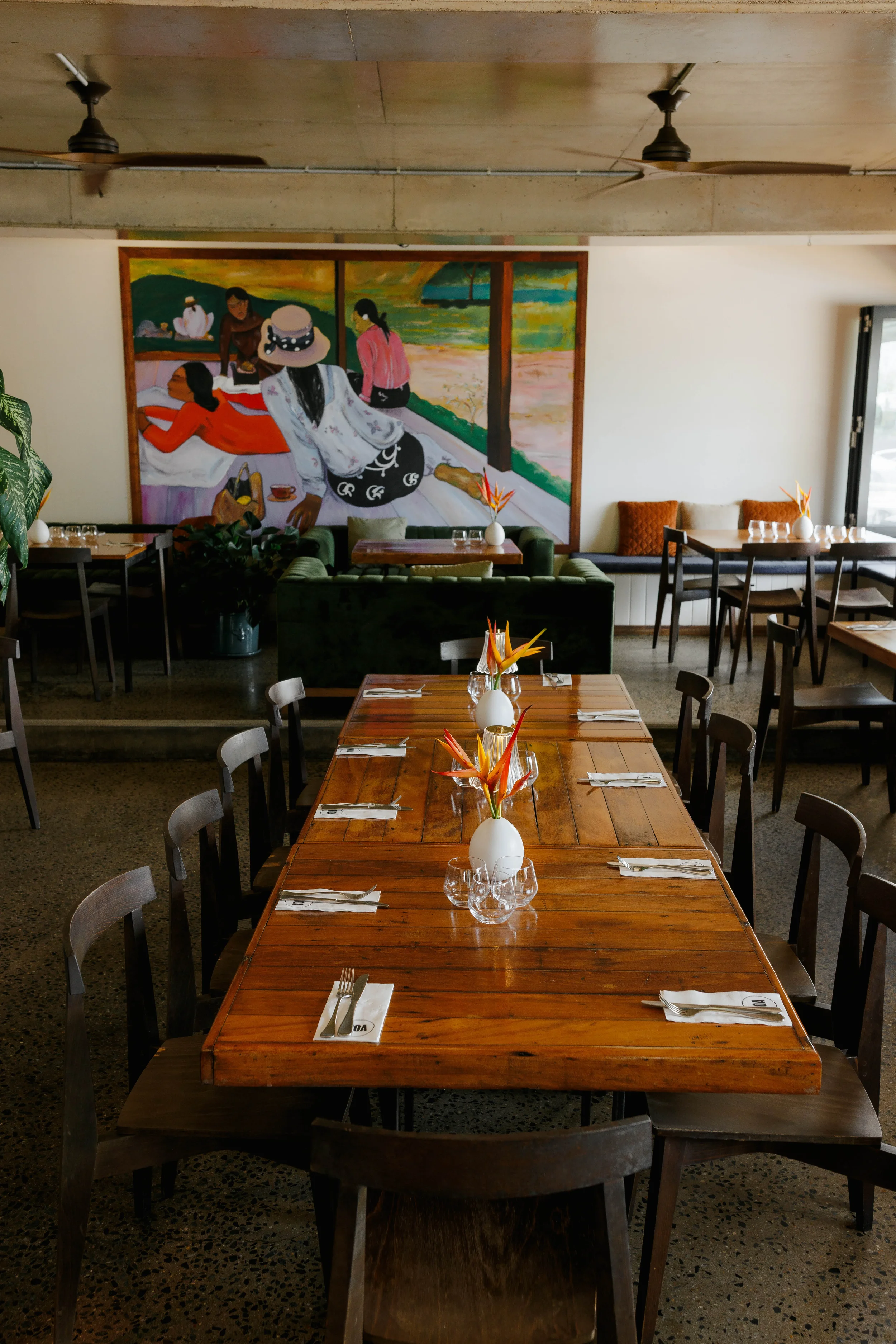A neatly set wooden dining table with floral centerpieces in a restaurant or cafe, with a colorful mural of people by a window on the wall in the background.