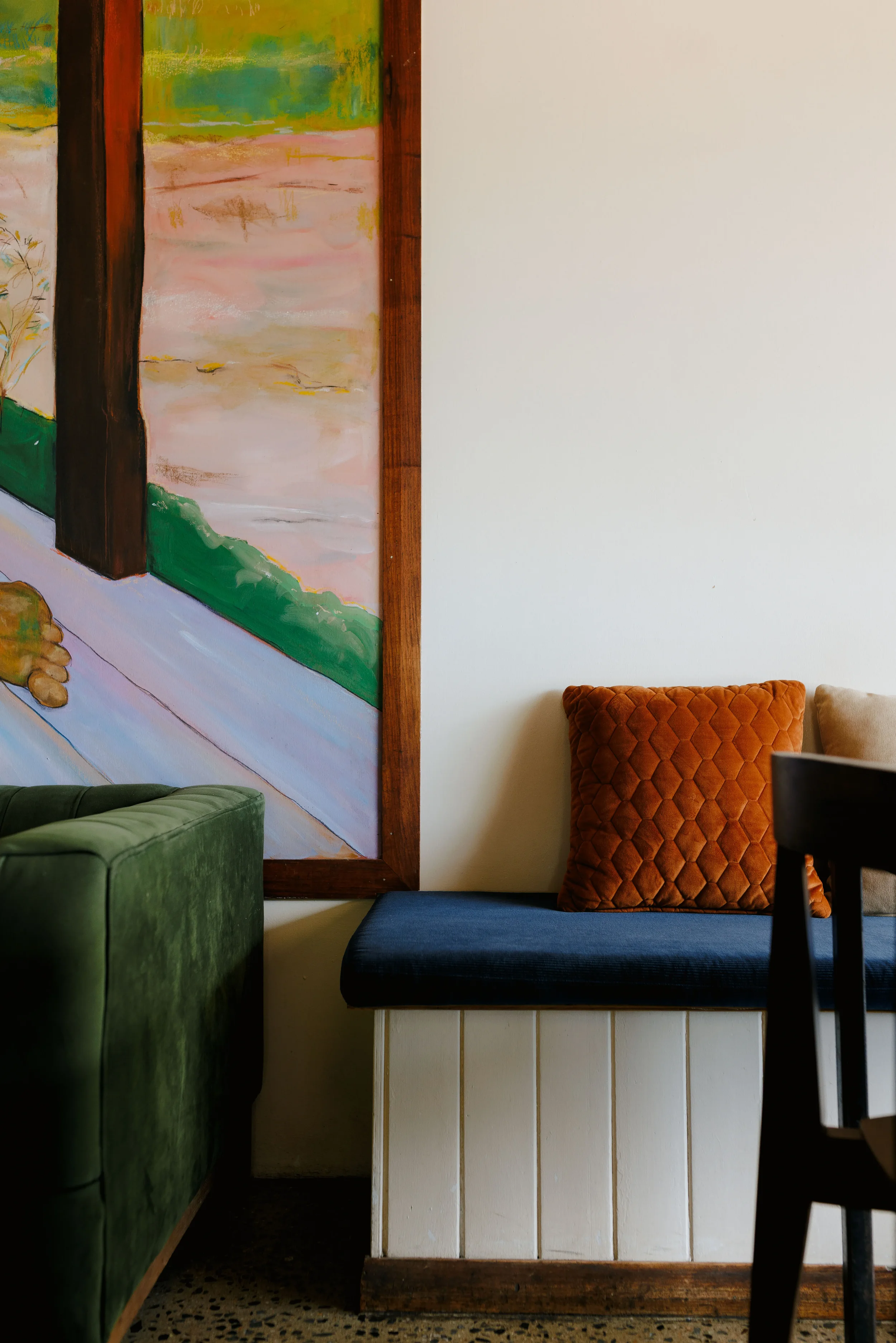 Interior scene with a white wall, colorful painting with abstract landscape, green armchair, and a seating area with an orange quilted pillow and navy blue cushion.
