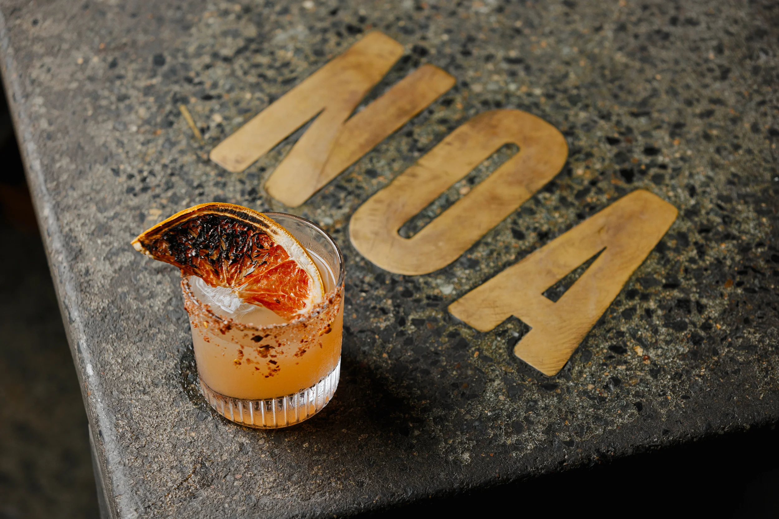 A cocktail in a short glass garnished with a grilled orange slice and chili powder rim, placed on a granite countertop with the word 'NOA' spelled out in wooden letters.