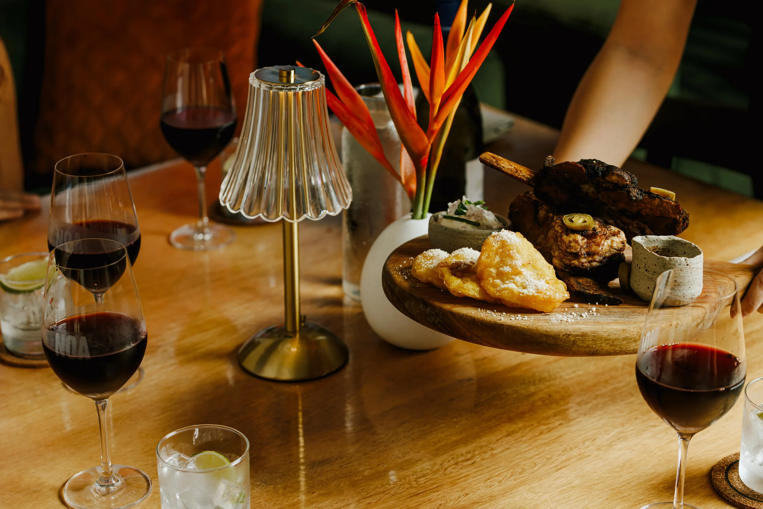 A wooden table set for a meal with multiple glasses of red wine, a white vase with orange and yellow flowers, and a round wooden platter with grilled meat, fried food, and a cup of sauce.