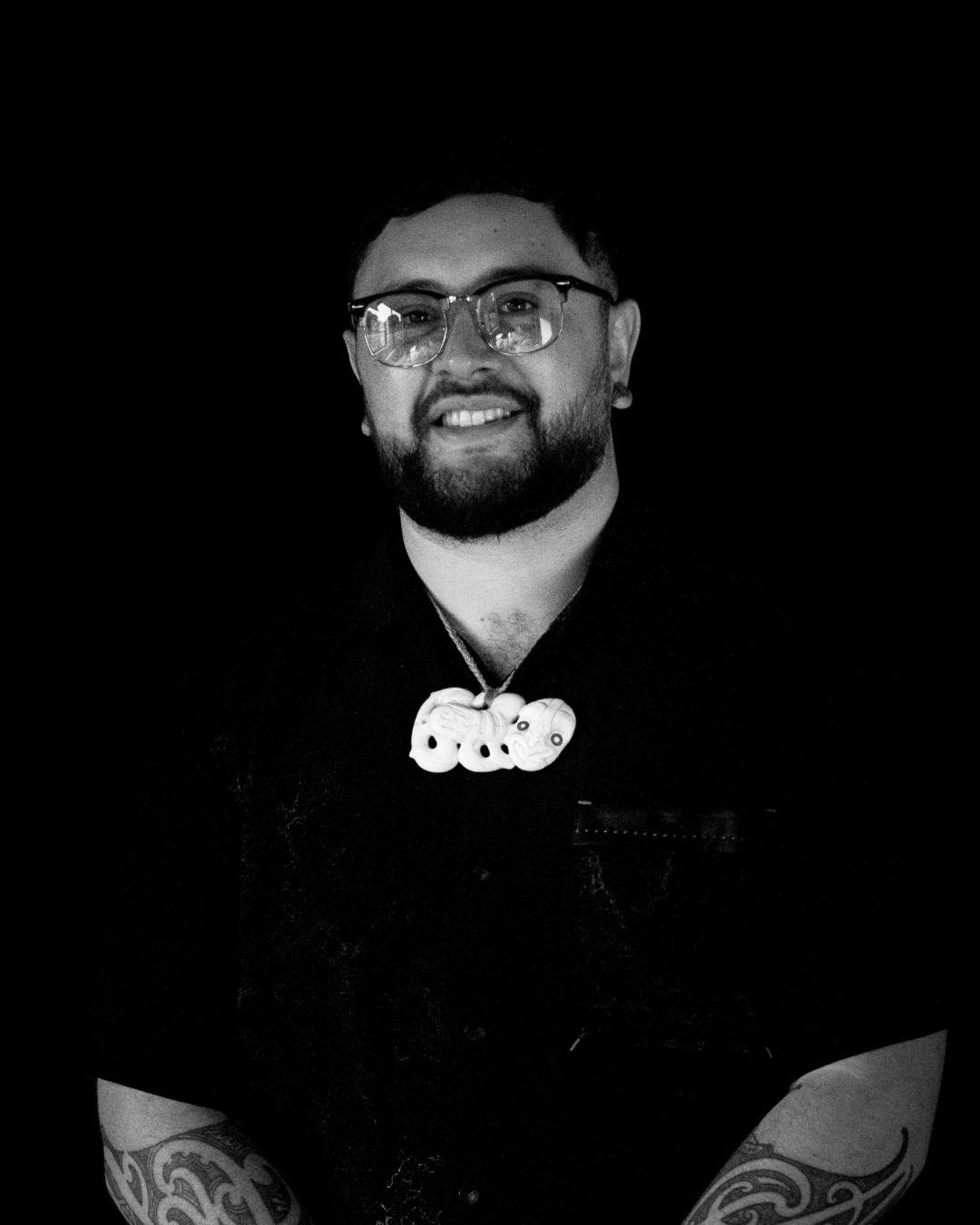 Black and white portrait of a man with glasses, a beard, and tattoos on his arms, wearing a necklace with large beads and a shirt with a name badge.