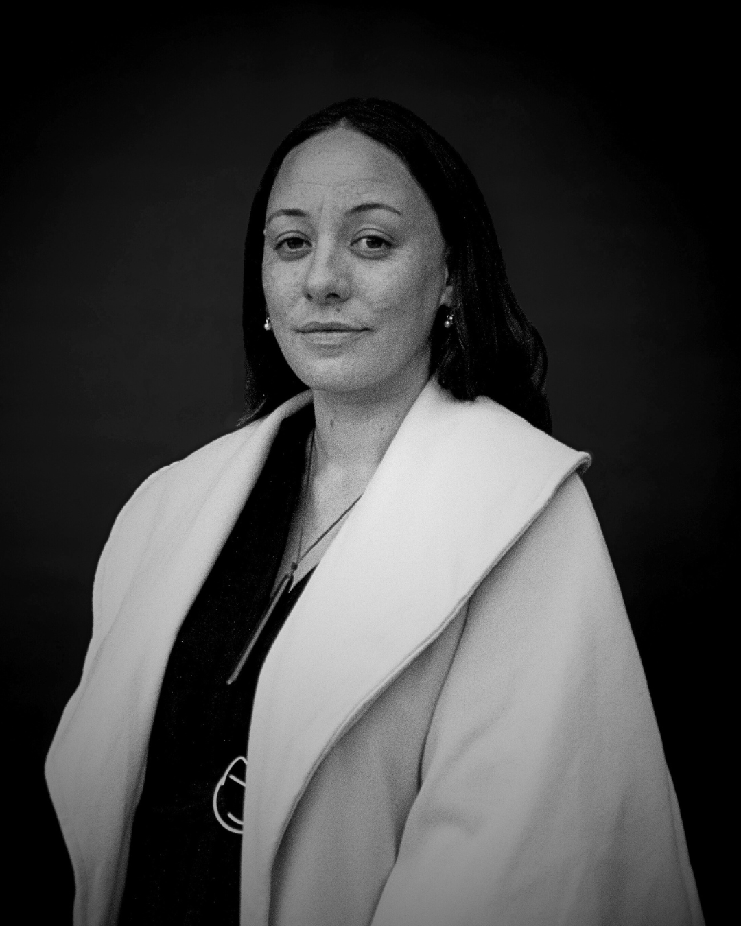 Black and white portrait of a woman with shoulder-length dark hair, wearing earrings, a dark top with a jacket or shawl over it, and a necklace, against a dark plain background.