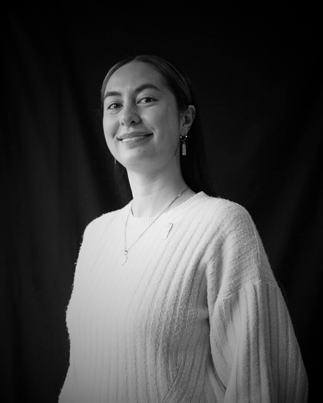 Black and white portrait of a smiling woman wearing a light-colored sweater, jewelry, and earrings, against a dark background.