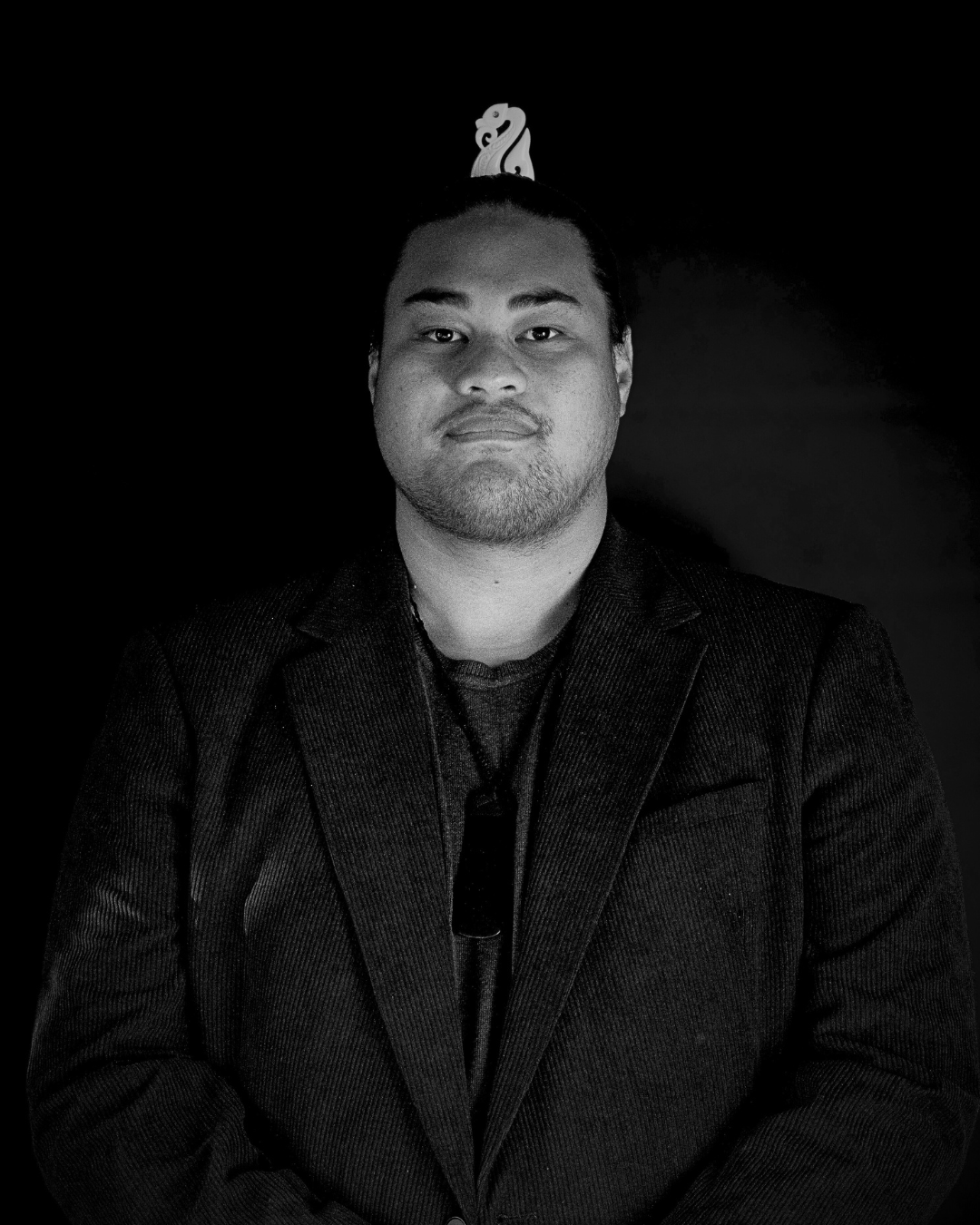 Black and white portrait of a young man with short hair, wearing a blazer, standing against a dark background with a decorative object above his head.