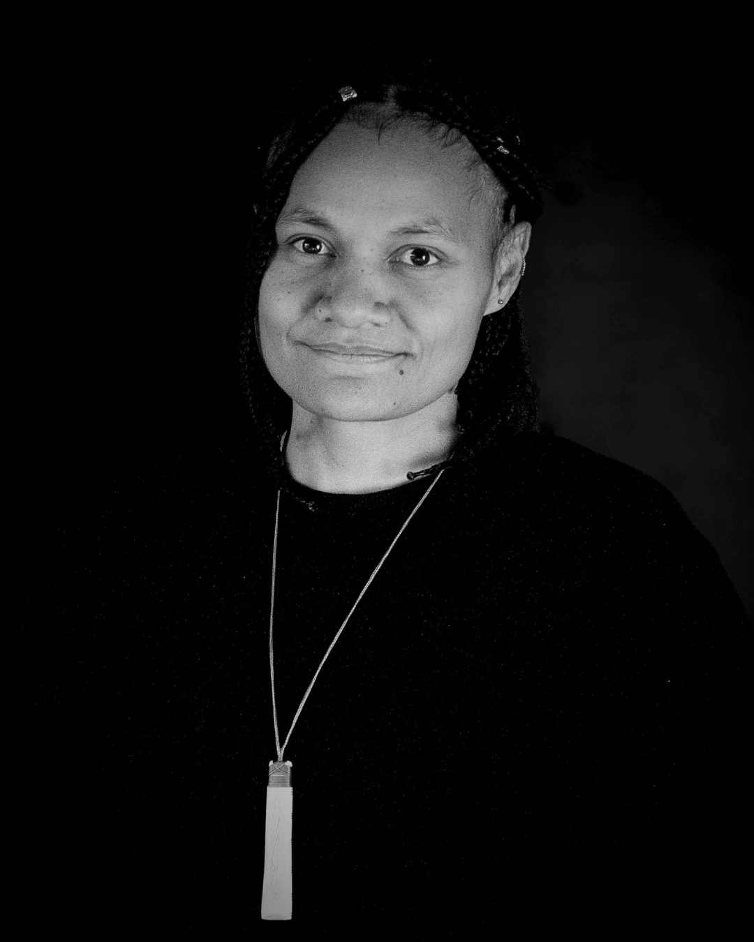 Black-and-white portrait of a woman with braids, smiling, wearing a necklace with a rectangular pendant.