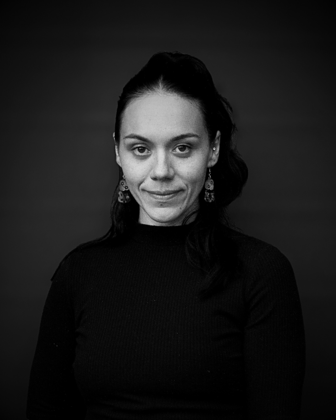 Black and white portrait of a woman with dark hair, wearing earrings and a black top, against a dark background.