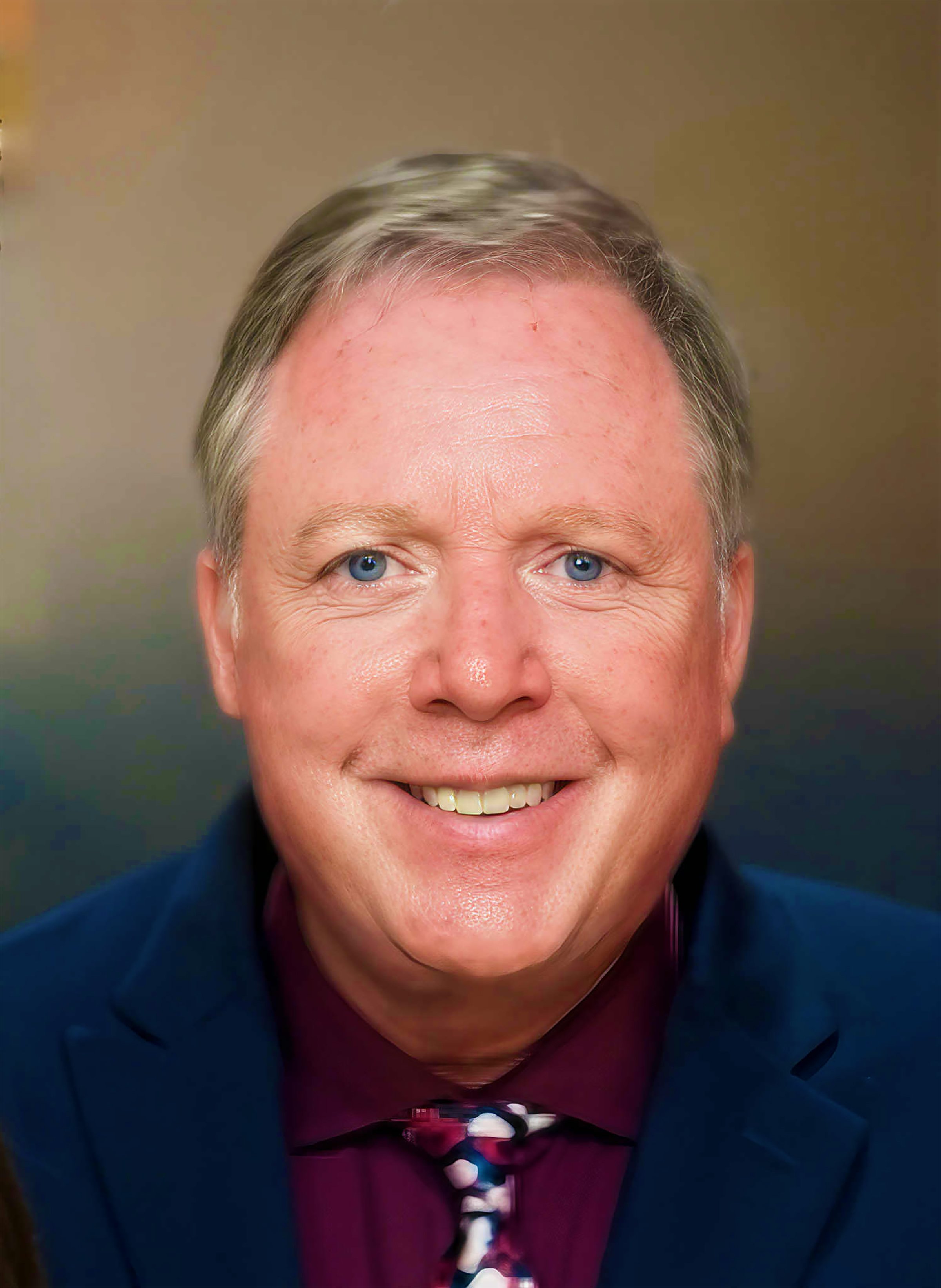 A smiling middle-aged man with gray hair and blue eyes, wearing a navy blazer, a maroon shirt, and a patterned tie, against a plain background.