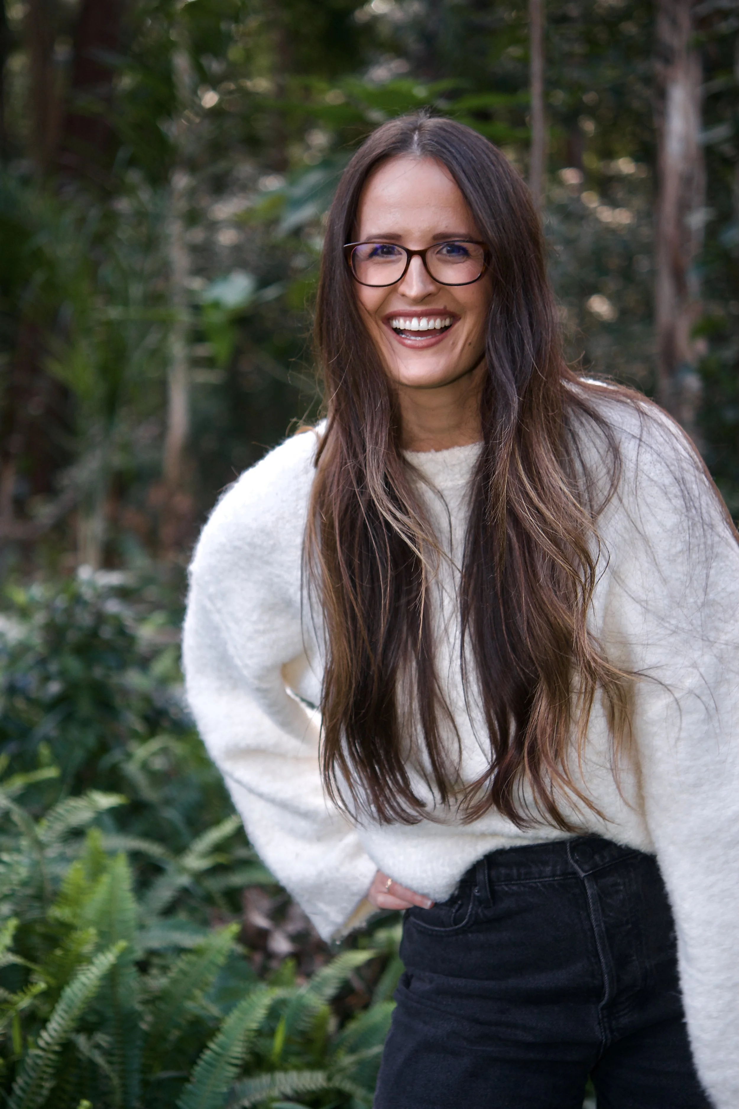 Headshot of Hayley Hughes, author of Thank You, Burnout: From depletion to peace smiling. Hayley wears black rimmed glasses and a white knit top against the green, nature-filled background.