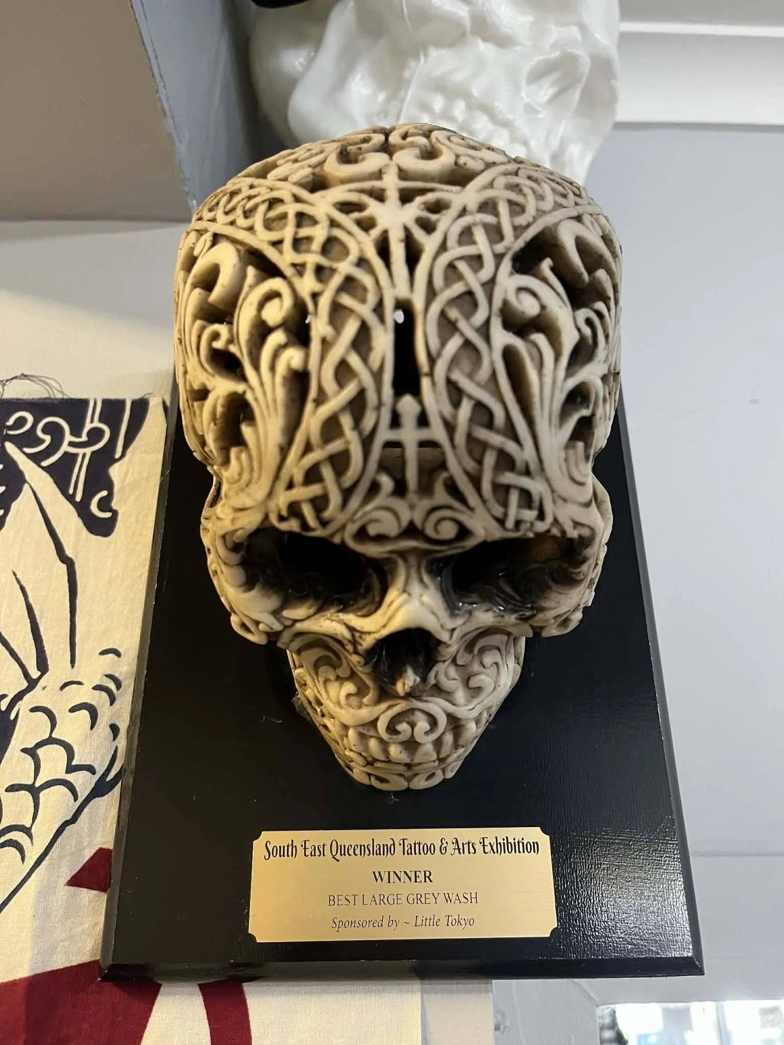 Decorative skull sculpture with intricate carved patterns, awarded as Best Large Grey Wash at the South East Queensland Tattoo & Arts Exhibition, placed on a black display stand.