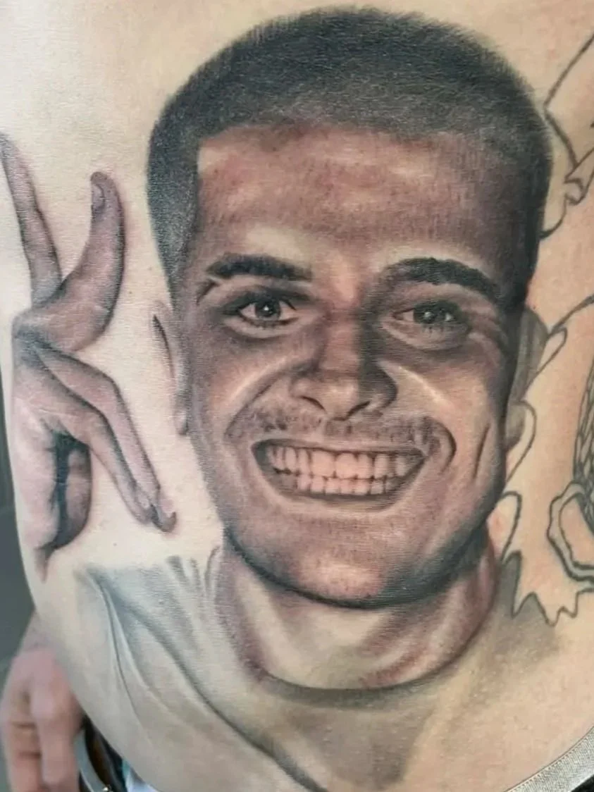 Tattoo of a smiling man's face making a peace sign with his hand.
