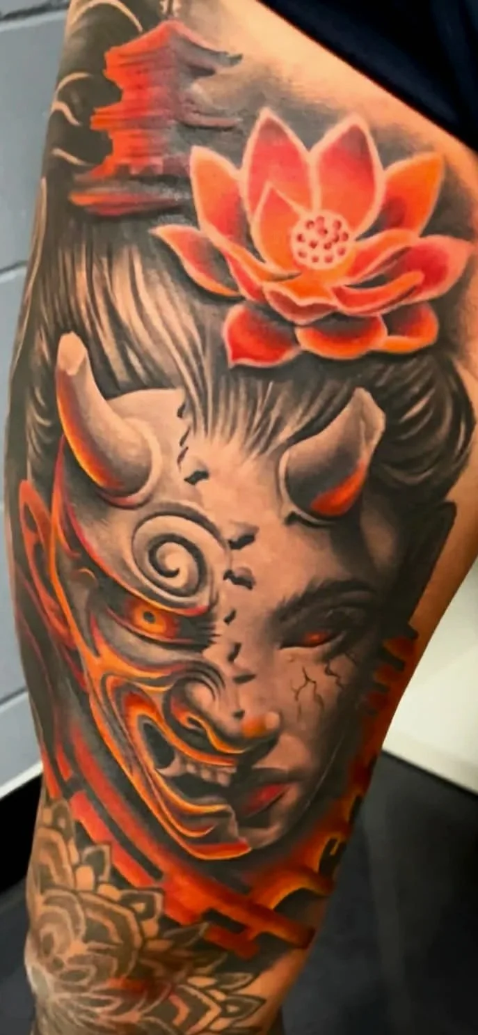 A colorful tattoo on a person's arm featuring a lotus flower, a woman's face with red eyes, horns, and swirling patterns in orange, black, and gray tones.