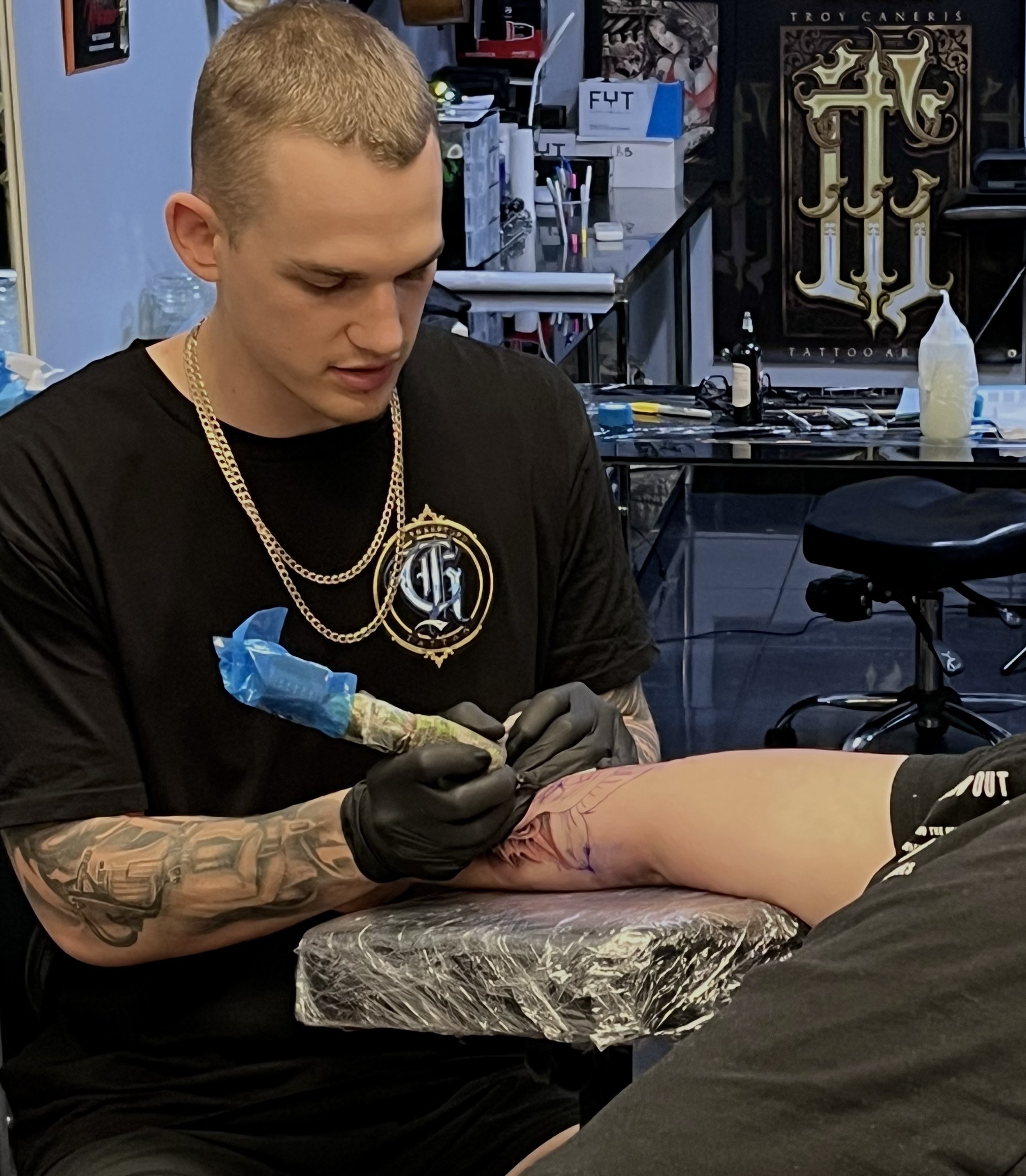 A tattoo artist tattooing a person's arm in a tattoo studio, with various equipment and artwork visible in the background.