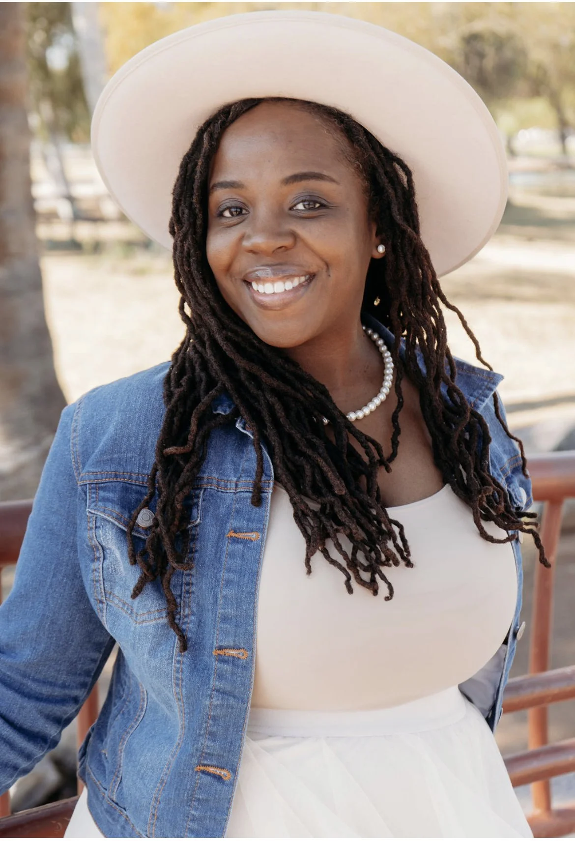 A woman with dark skin, long dreadlocks, wearing a white hat, pearl earrings and necklace, blue denim jacket, and white dress, smiling outdoors.
