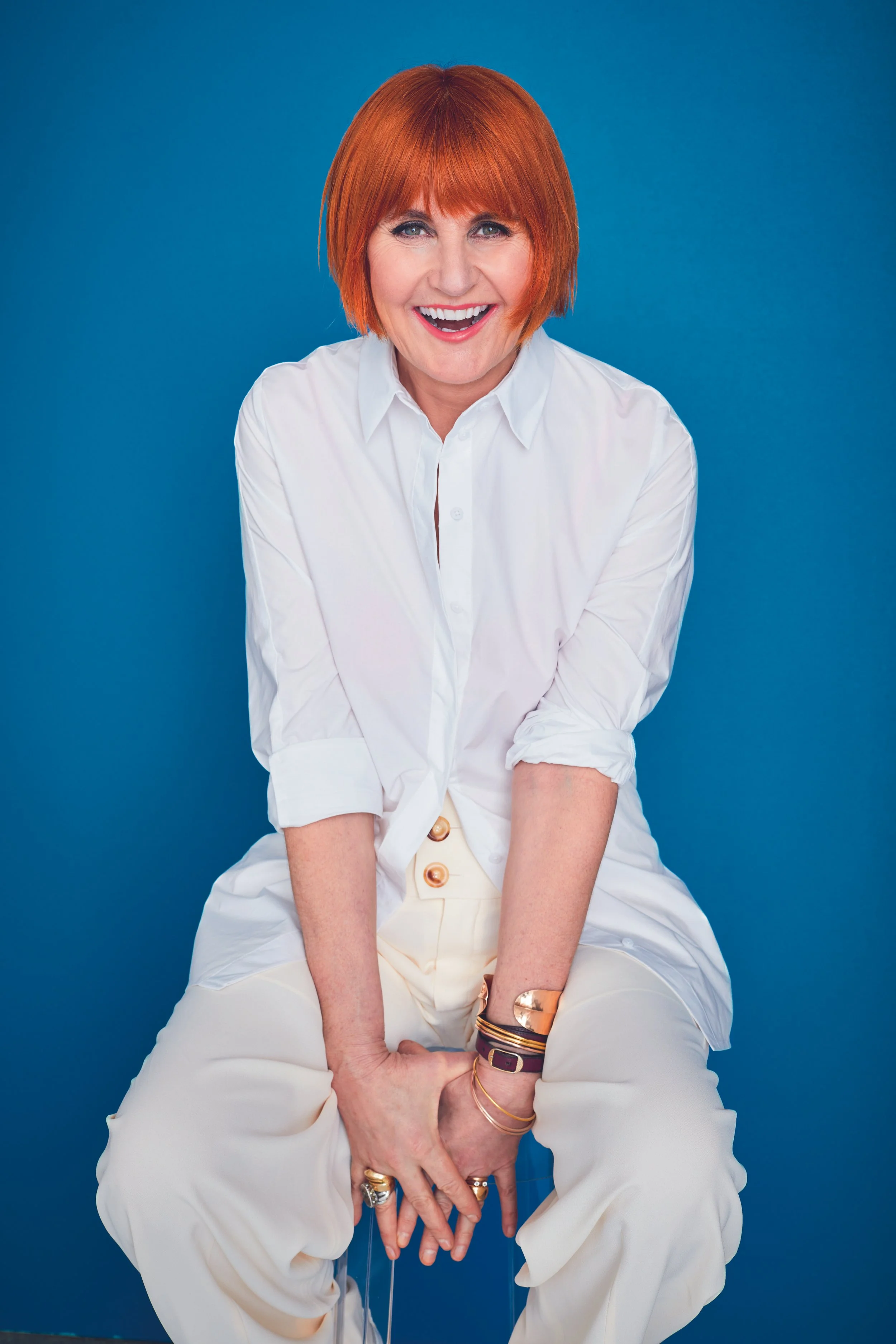 Smiling woman with red hair in a white blouse and cream-colored pants sitting against a blue background.